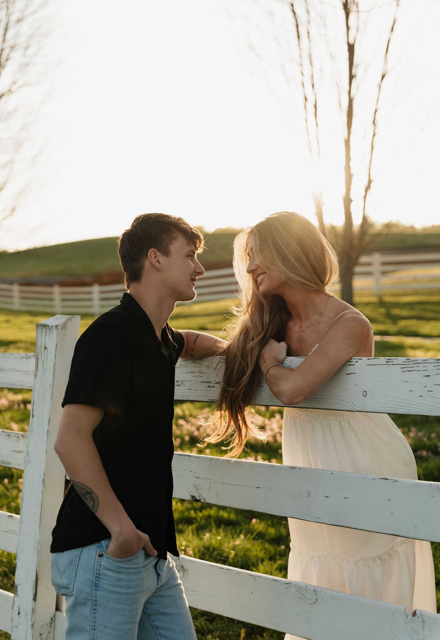 A romantic couple shares tender moments by a white wooden fence at sunset in a rural setting.