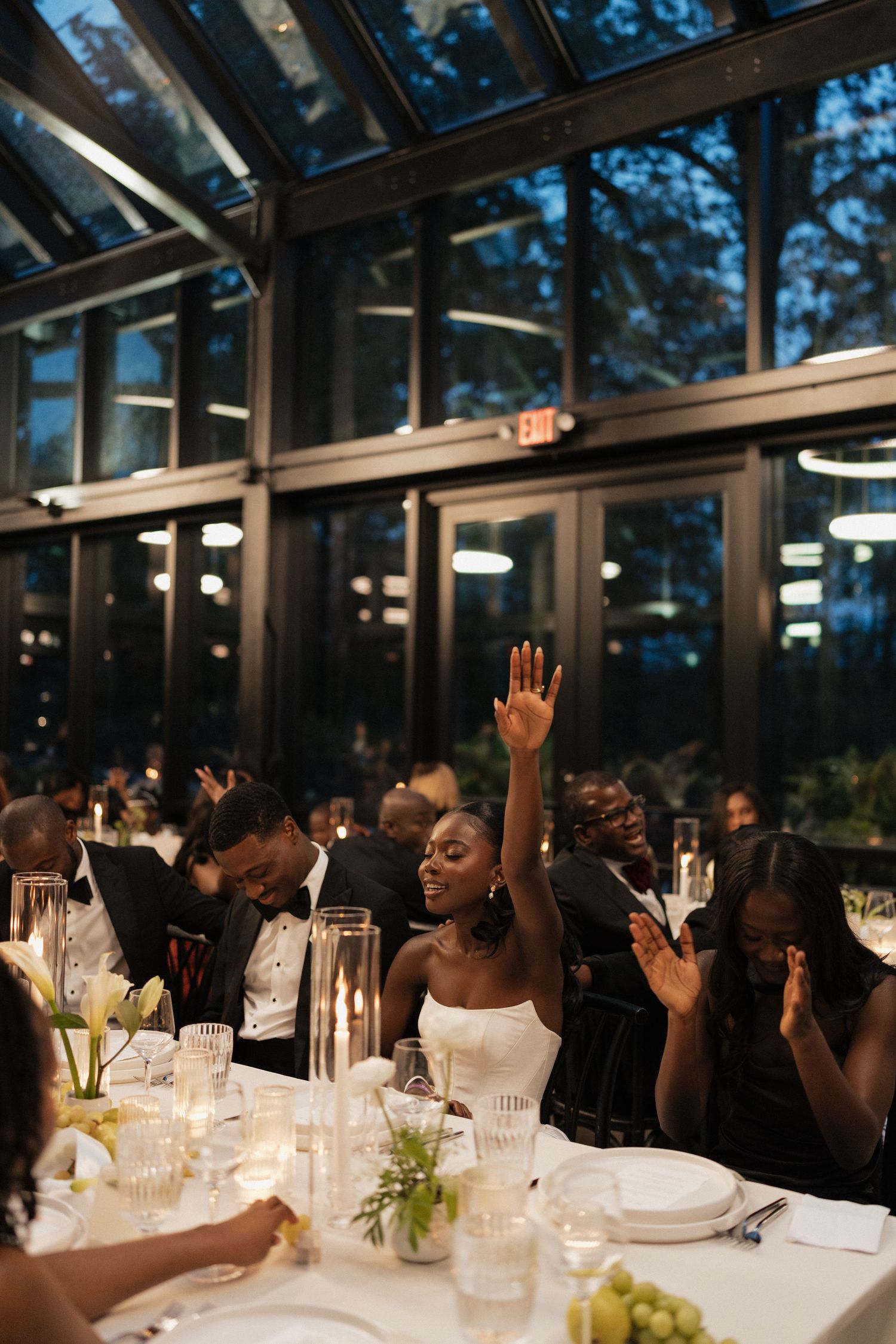 Wedding guests raise their hands in celebration during an elegant candlelit dinner reception in a glass-enclosed venue.