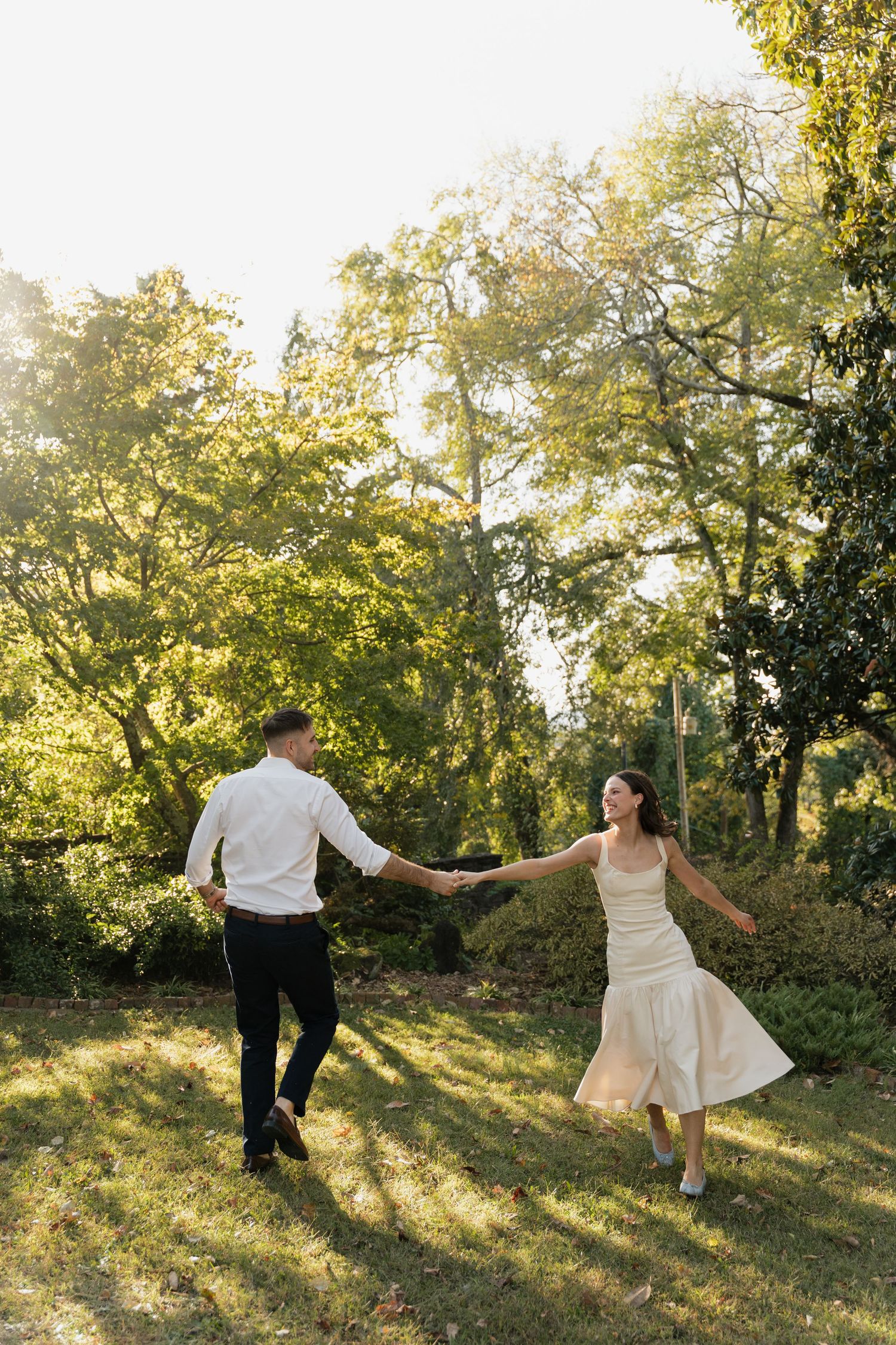 A couple in wedding attire dances together on a sunlit hillside surrounded by trees during golden hour.