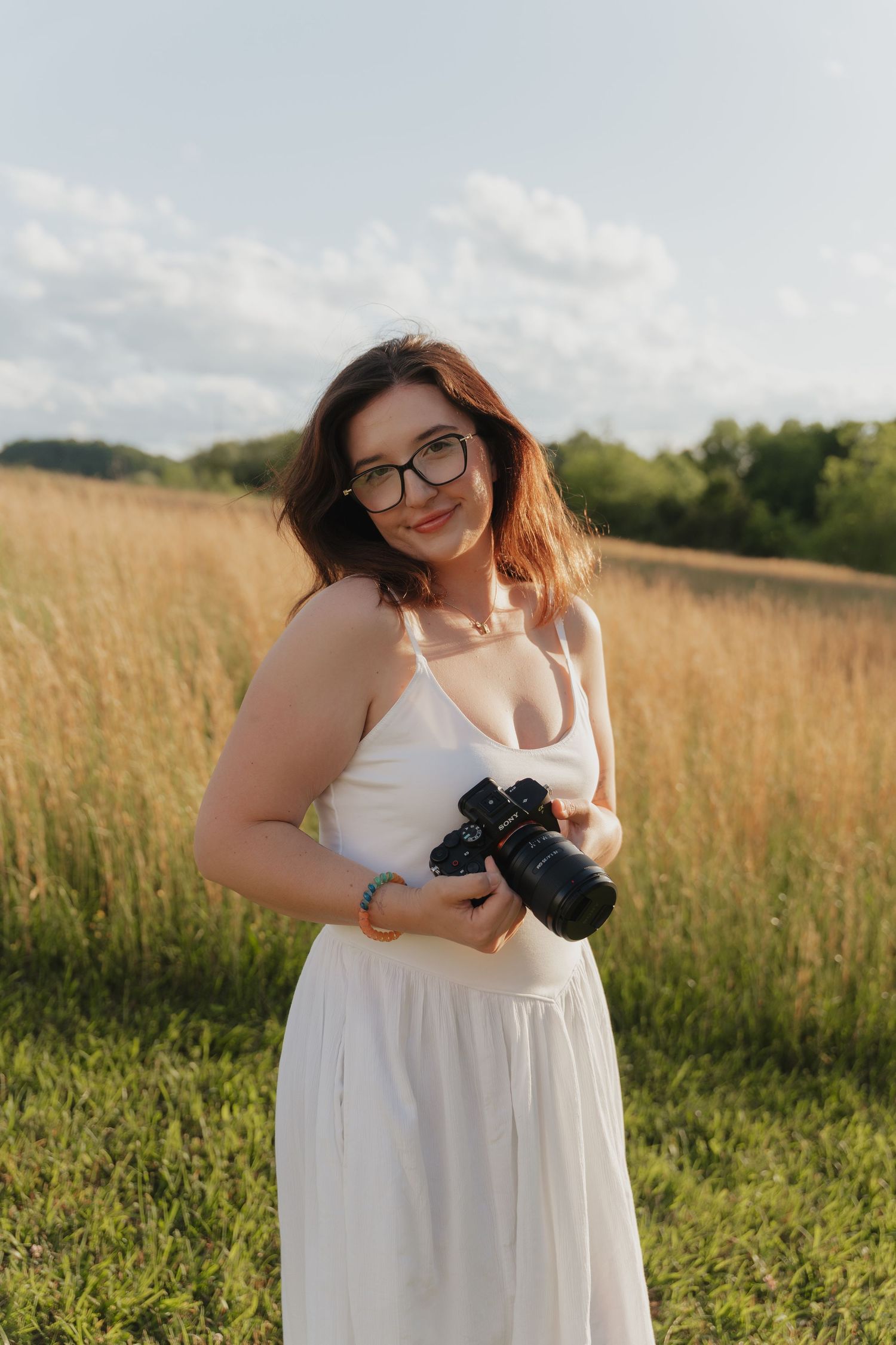 Photographer in a flowing white dress smiles while holding a camera in a sunlit meadow of tall grass.