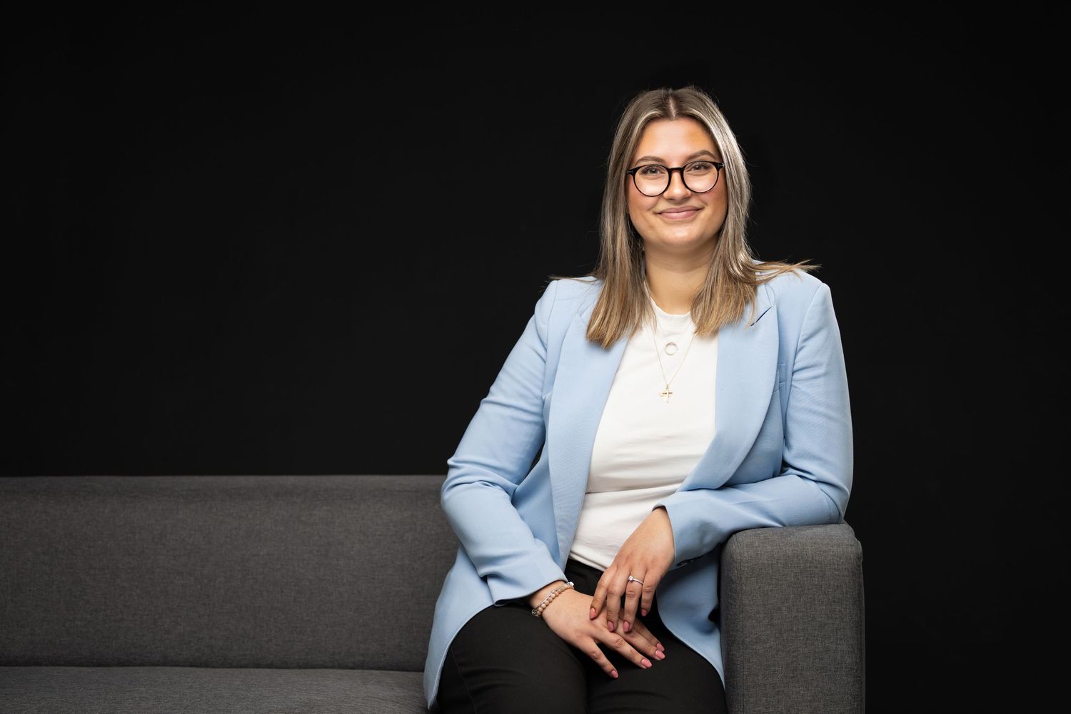 Professional woman in light blue blazer and glasses sitting on gray sofa against dark background.