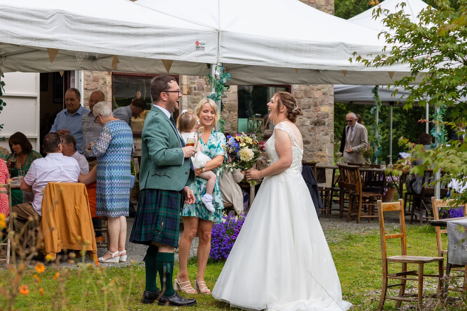 Wedding guests gather outdoors under white tents for a celebration in a garden setting.