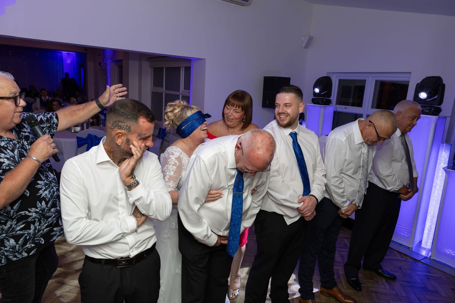 Group of guests dancing and laughing together at a wedding reception in a decorated venue with blue lighting.