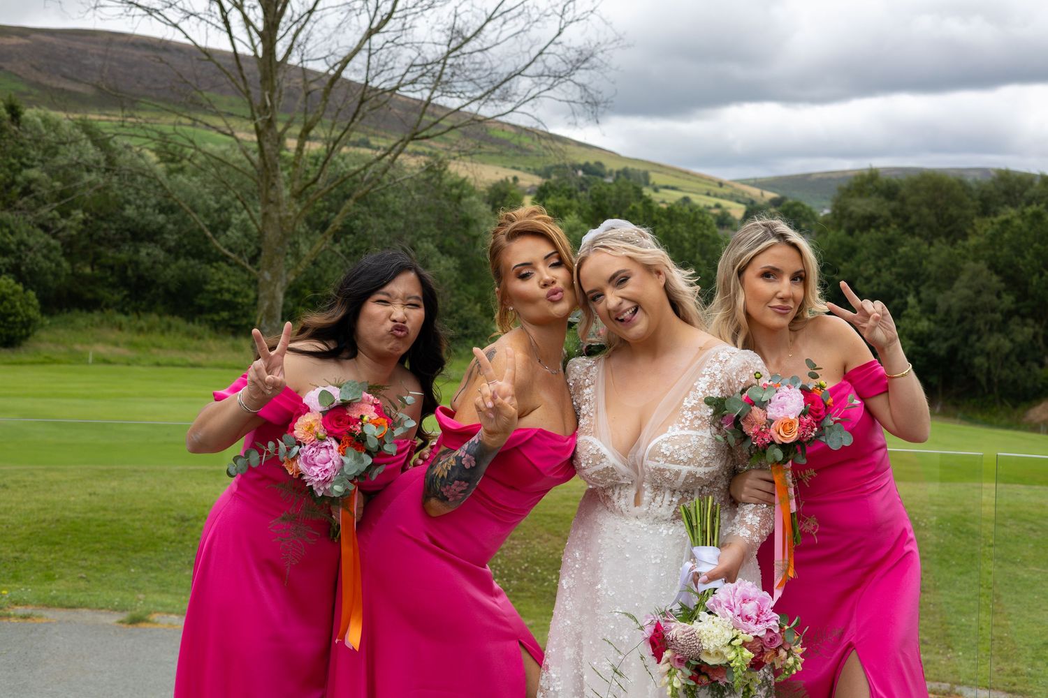 Bride in white lace gown poses with three bridesmaids in hot pink dresses holding bouquets on a green hillside.