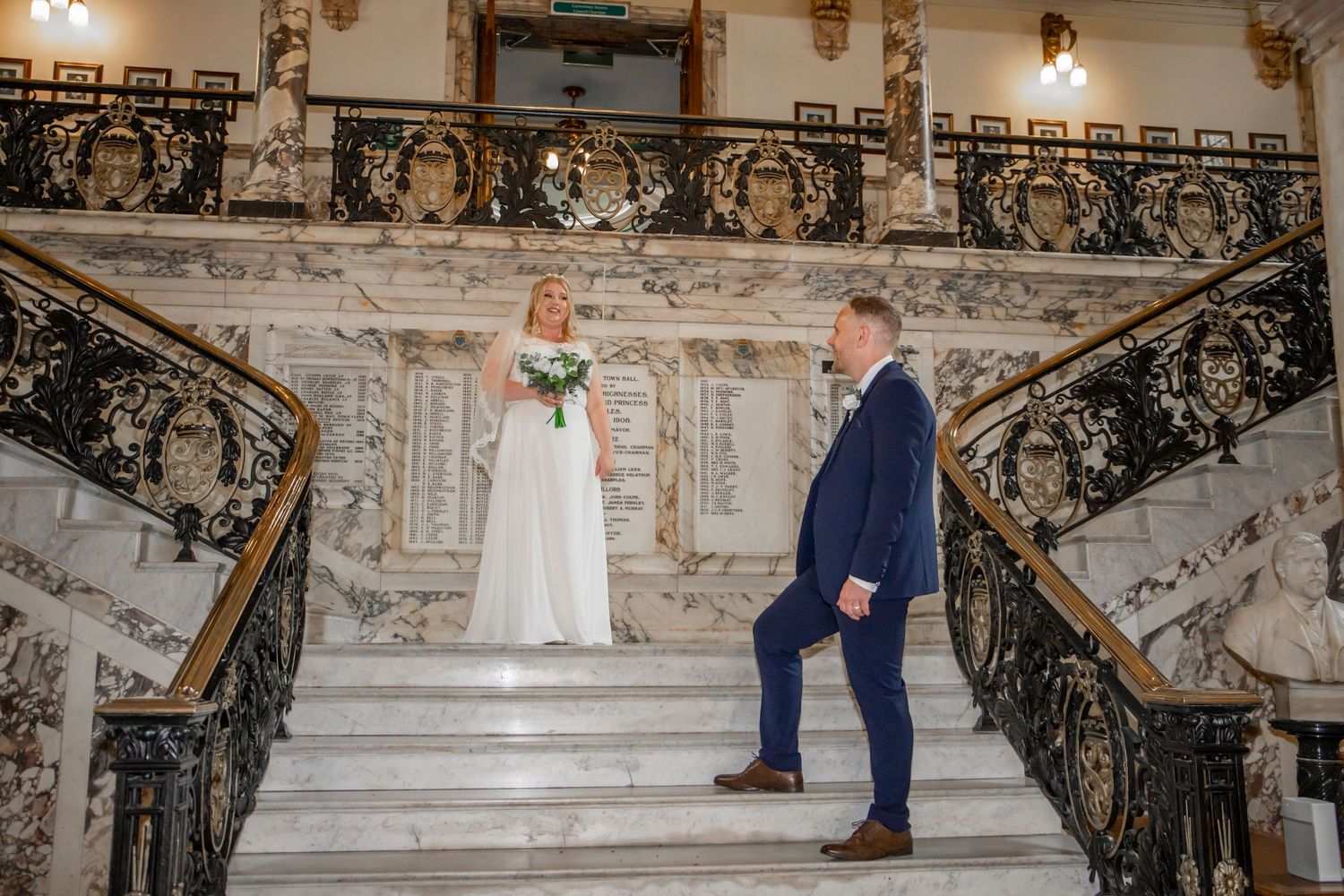 Bride and groom pose elegantly on ornate marble staircase with intricate wrought iron railings.