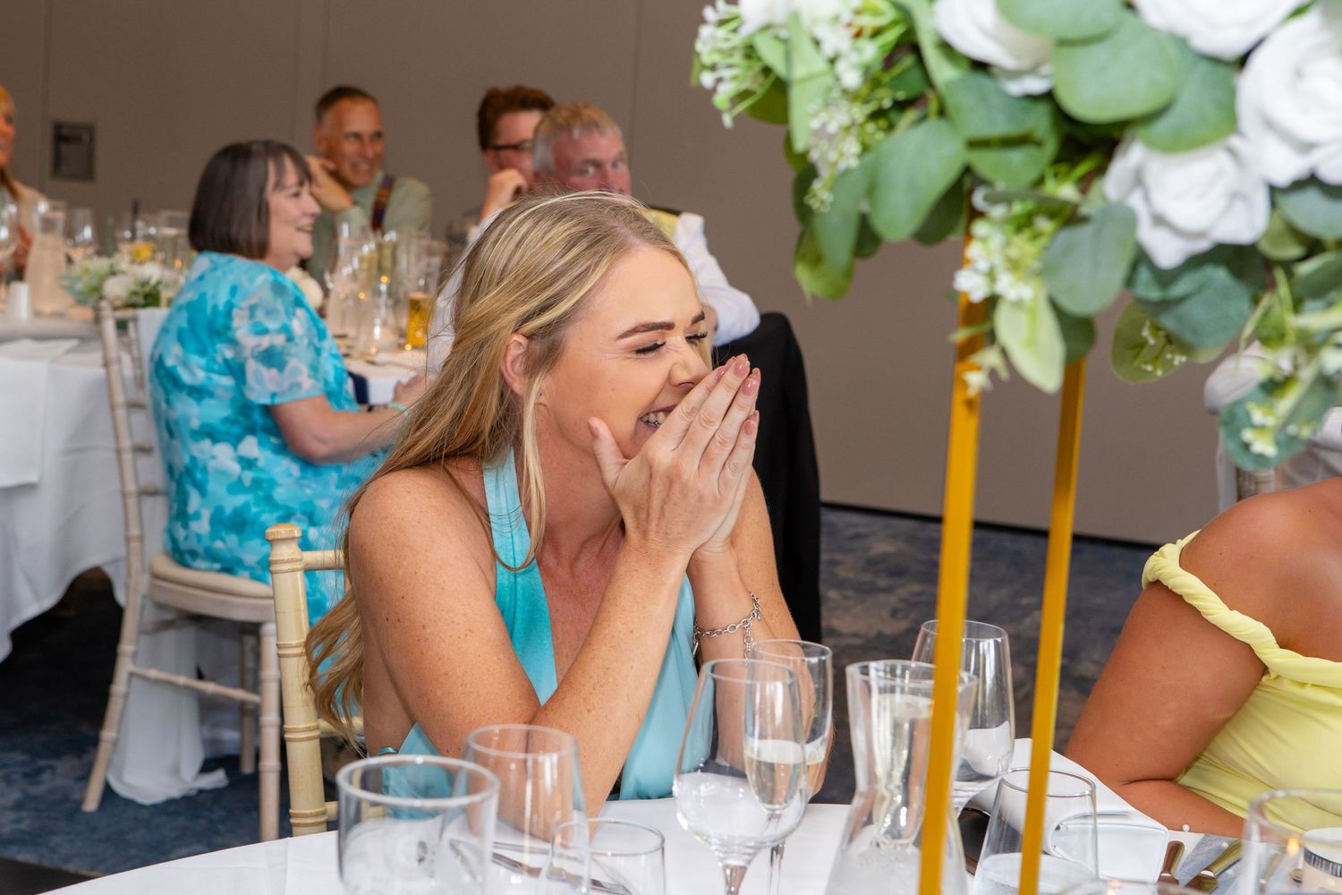 Guest in turquoise dress laughs with hand covering mouth at elegant dinner table with floral centerpiece.