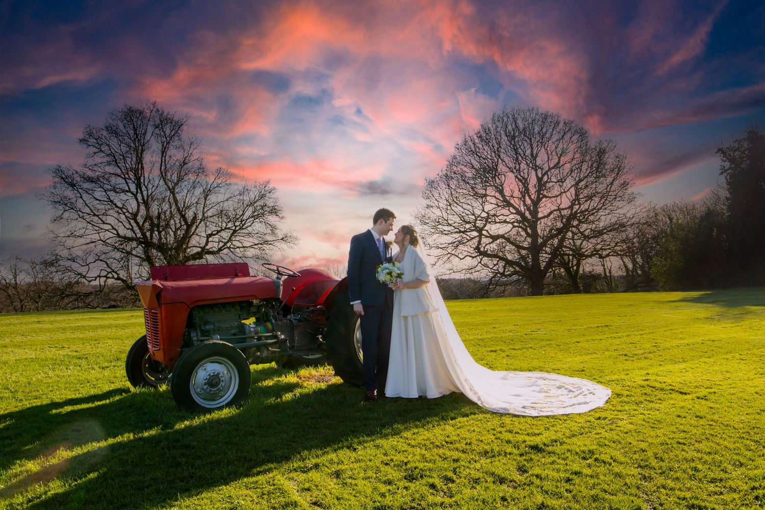 A romantic countryside wedding photo at sunset featuring a vintage red tractor in a green field.