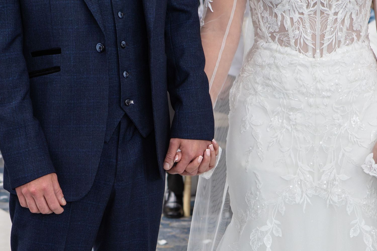 A couple in formal wedding attire stands together, with the bride wearing a white lace gown and the groom in a navy suit.