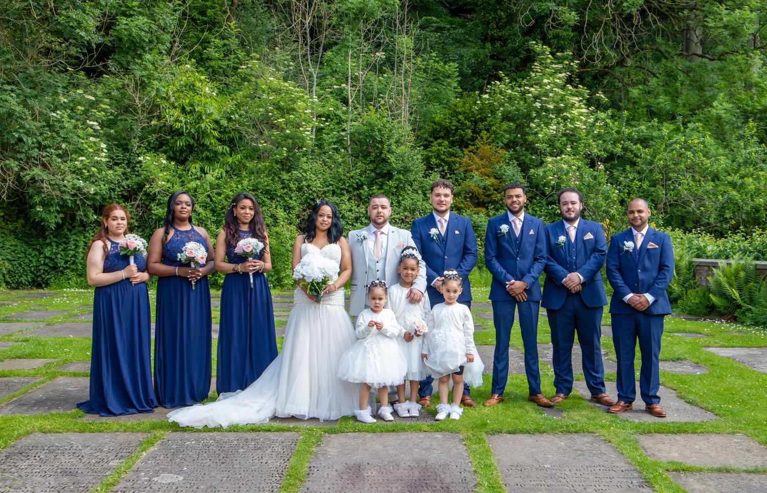 Wedding party lineup featuring bridesmaids in navy dresses and groomsmen in blue suits pose outdoors.