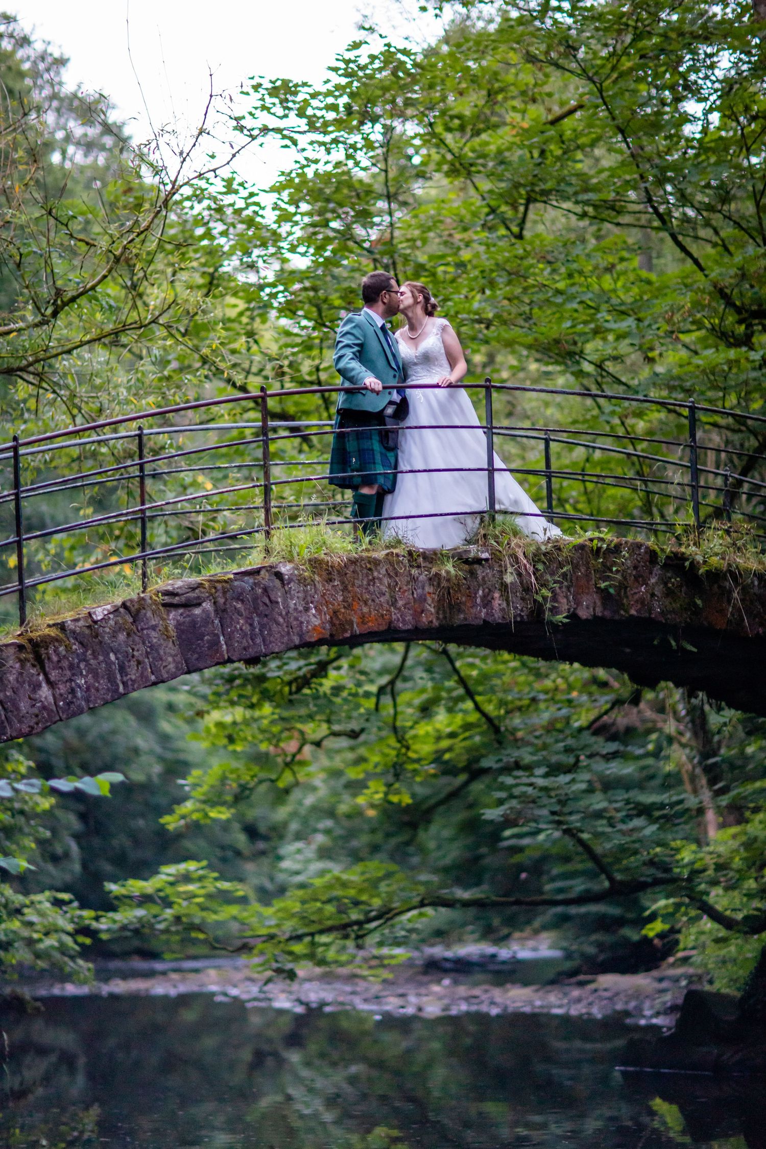 Wedding couple sharing a romantic moment on a rustic stone bridge surrounded by lush greenery.