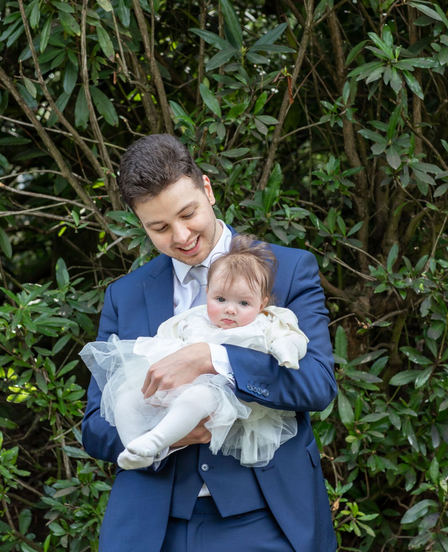 A tender moment between parent and baby in formal attire against green foliage.