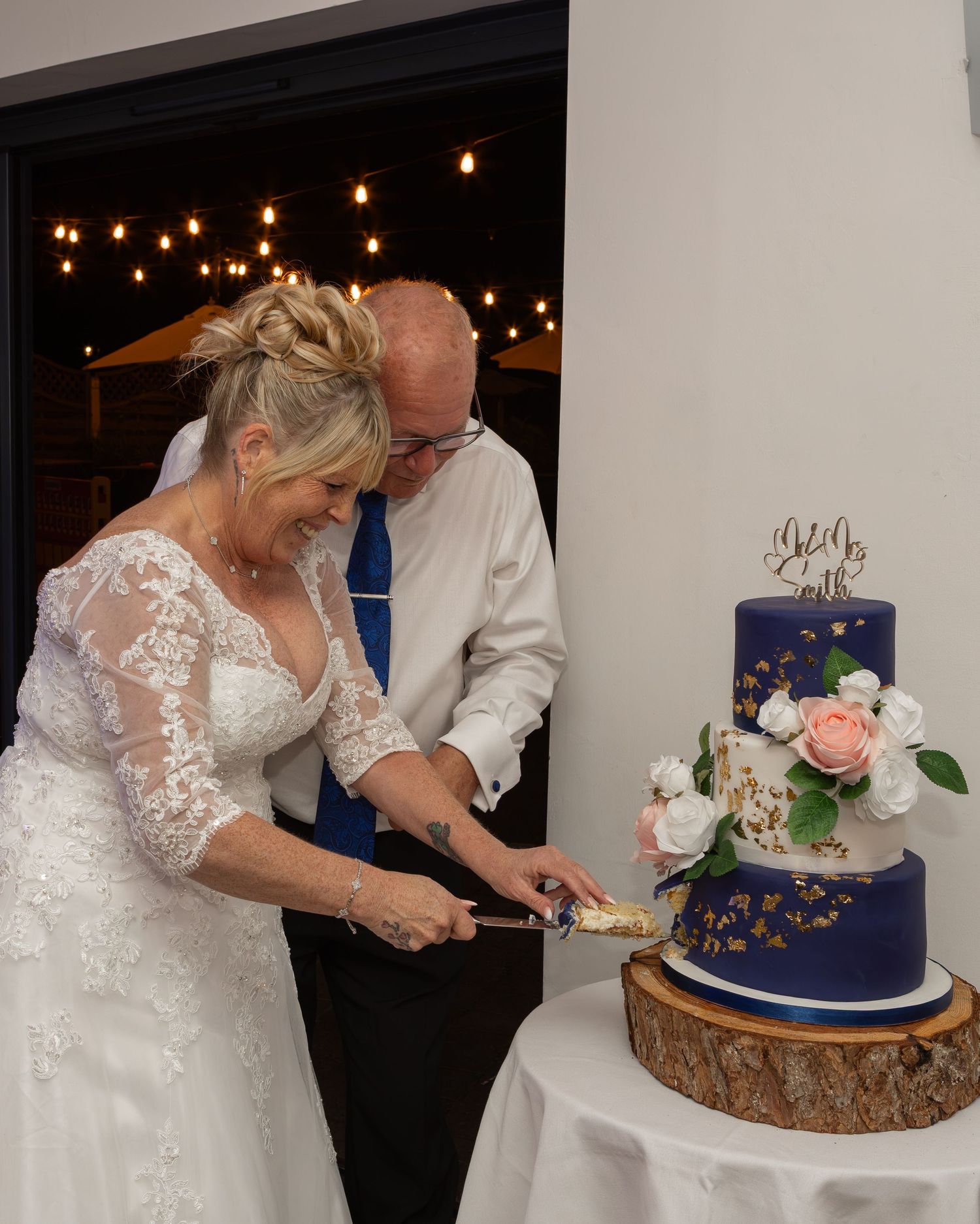 Bride and groom cutting a navy blue tiered wedding cake decorated with flowers on a rustic wood slice stand.