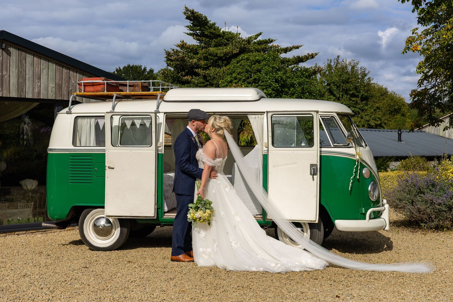 Bride and groom kissing beside a vintage green and white VW camper van at an outdoor wedding venue.