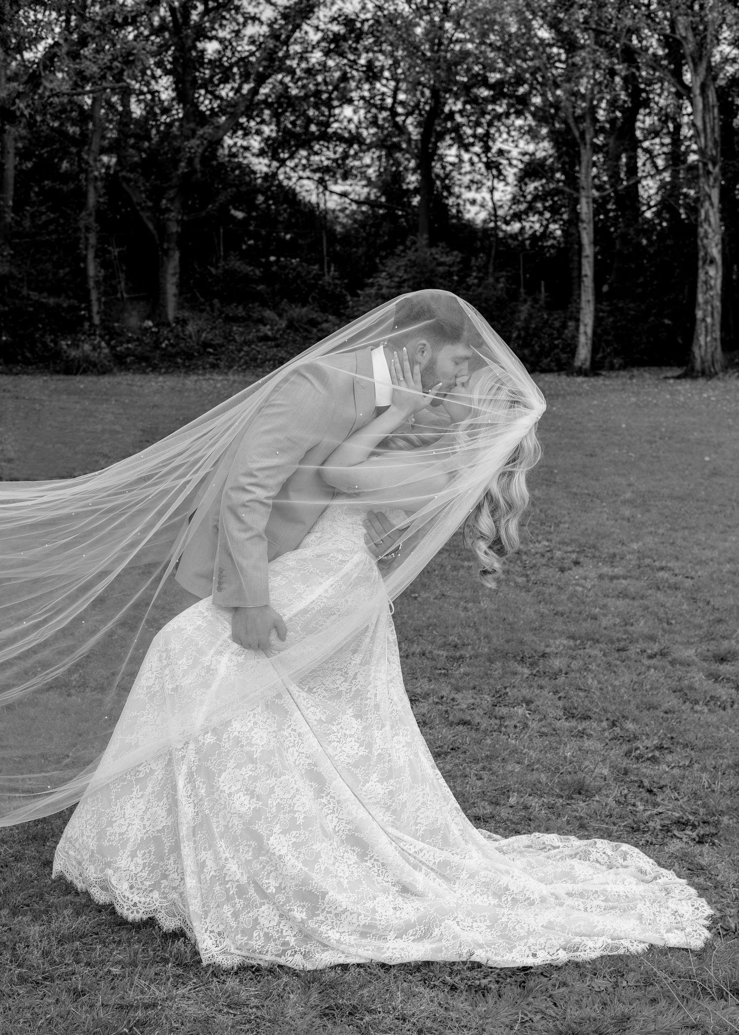 Black and white photo of bride and groom sharing a kiss under flowing veil on green lawn with trees.