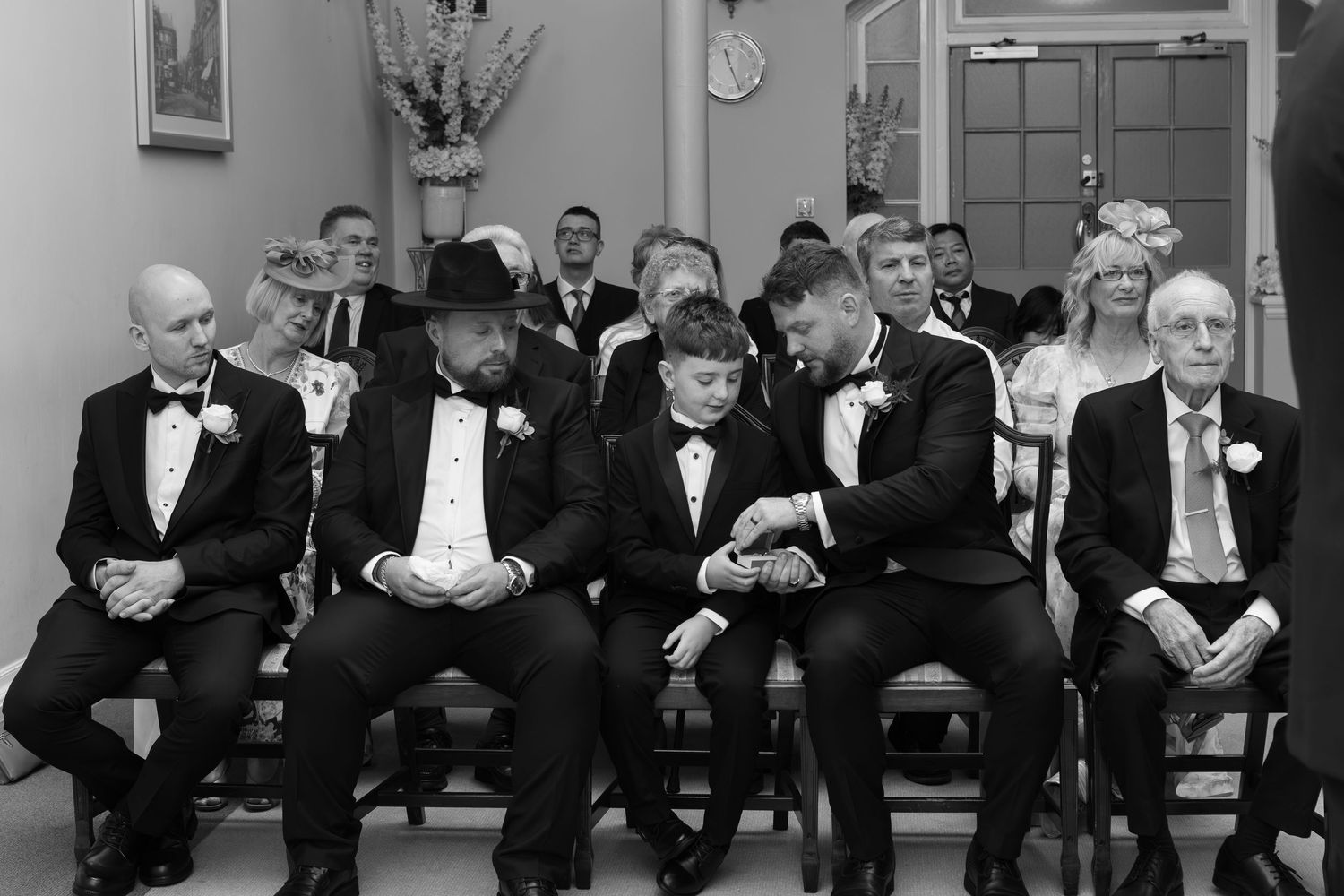 Black and white wedding photo of groomsmen seated together in formal attire during a ceremony.