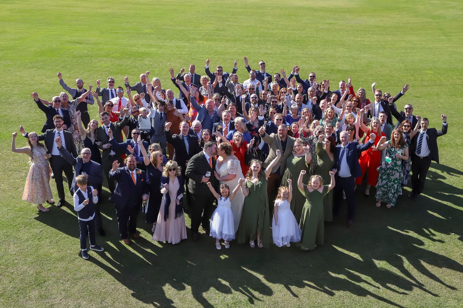 Aerial view of large wedding party group photo with guests arranged in a heart shape formation on green lawn.