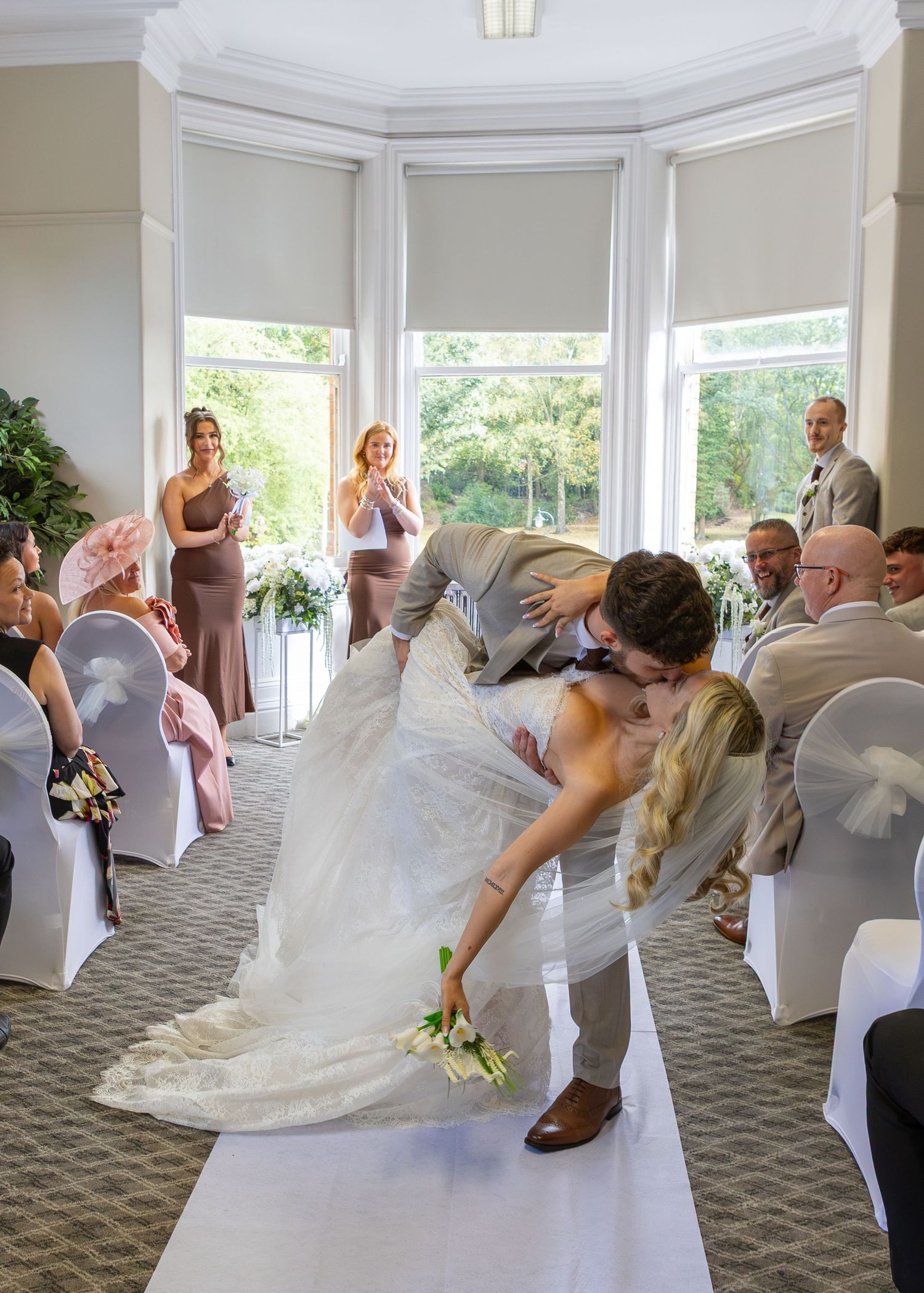 Bride and groom share a romantic dip kiss at wedding ceremony aisle with bridesmaids and guests applauding in background.