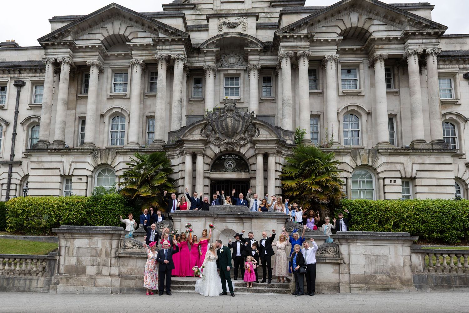 Wedding party posing on steps of grand neoclassical building with bride in pink gown.