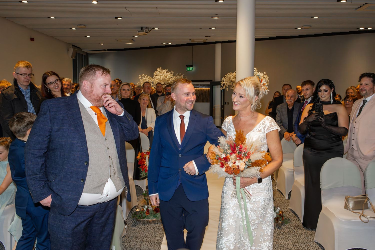 Wedding guests gather in a modern venue space with guests wearing formal attire including suits and evening wear.