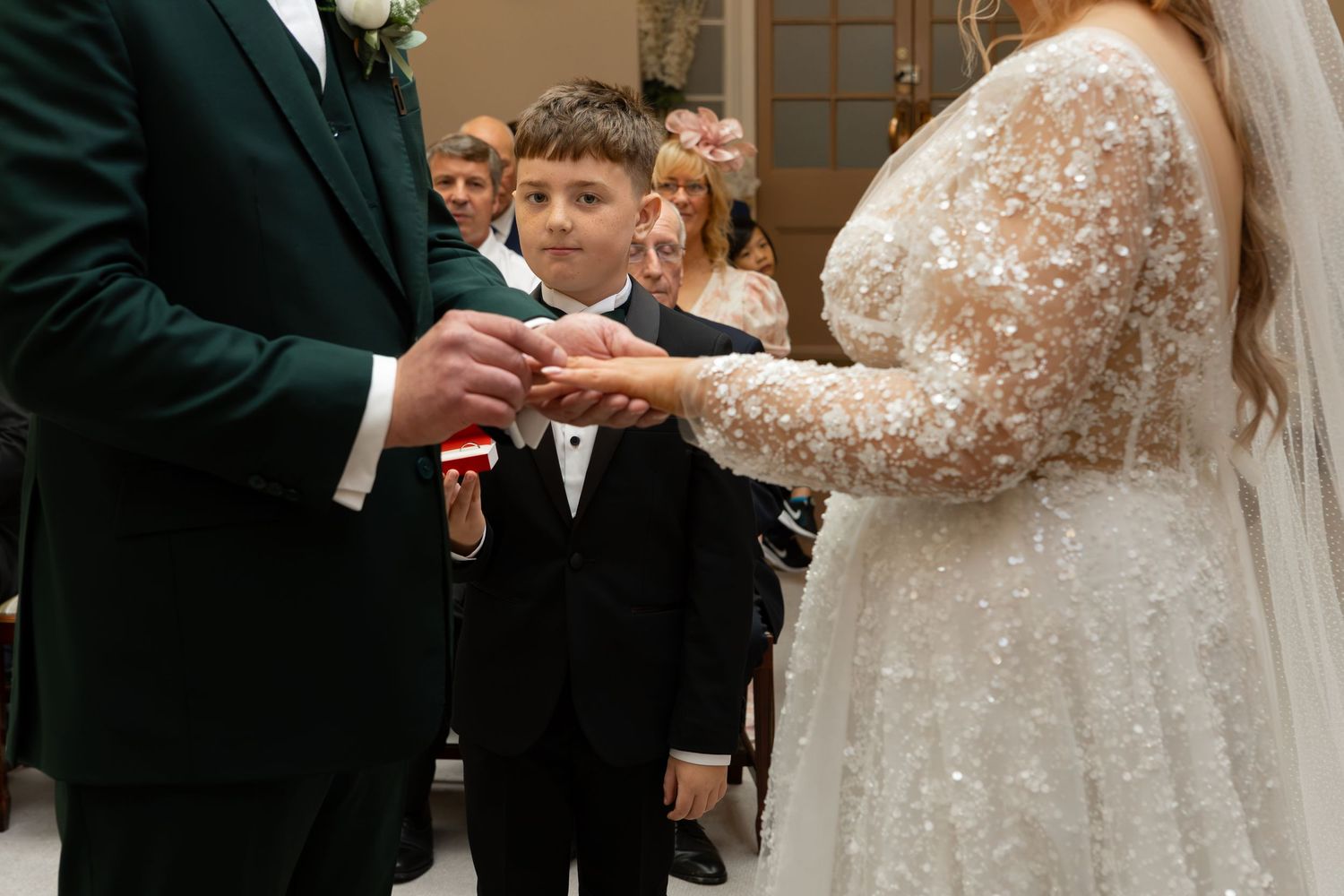 A young boy in a tuxedo watches a couple exchange wedding rings during a ceremony.