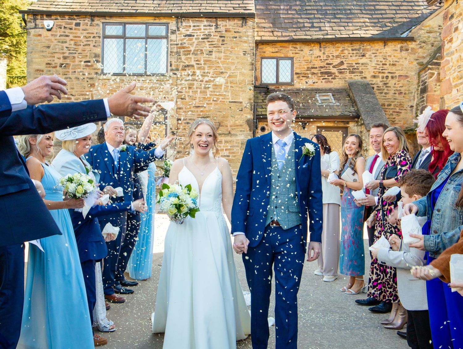 Wedding celebration outside stone building with guests throwing confetti.