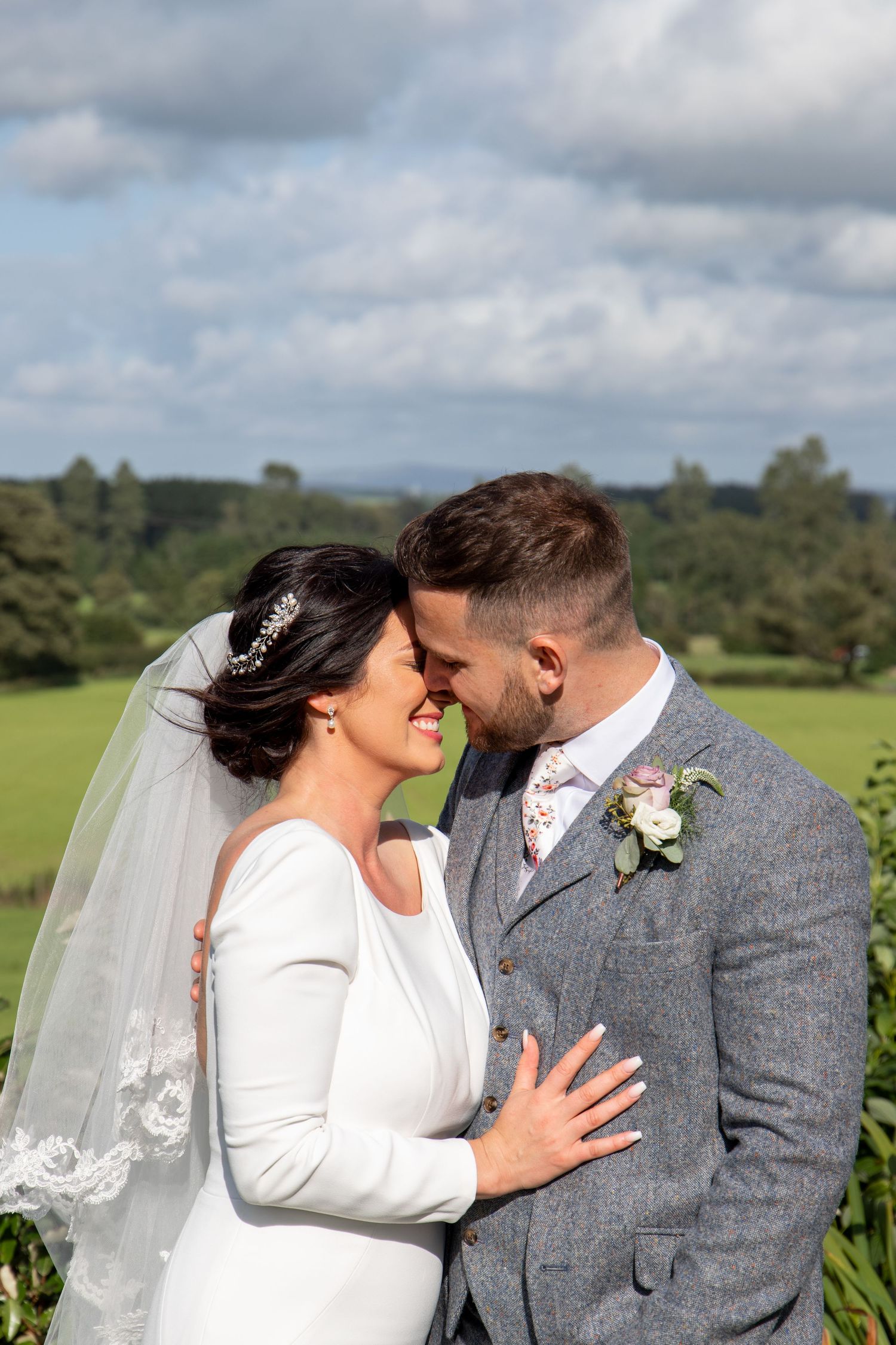Wedding couple embraces while laughing together in a scenic countryside setting with cloudy skies.