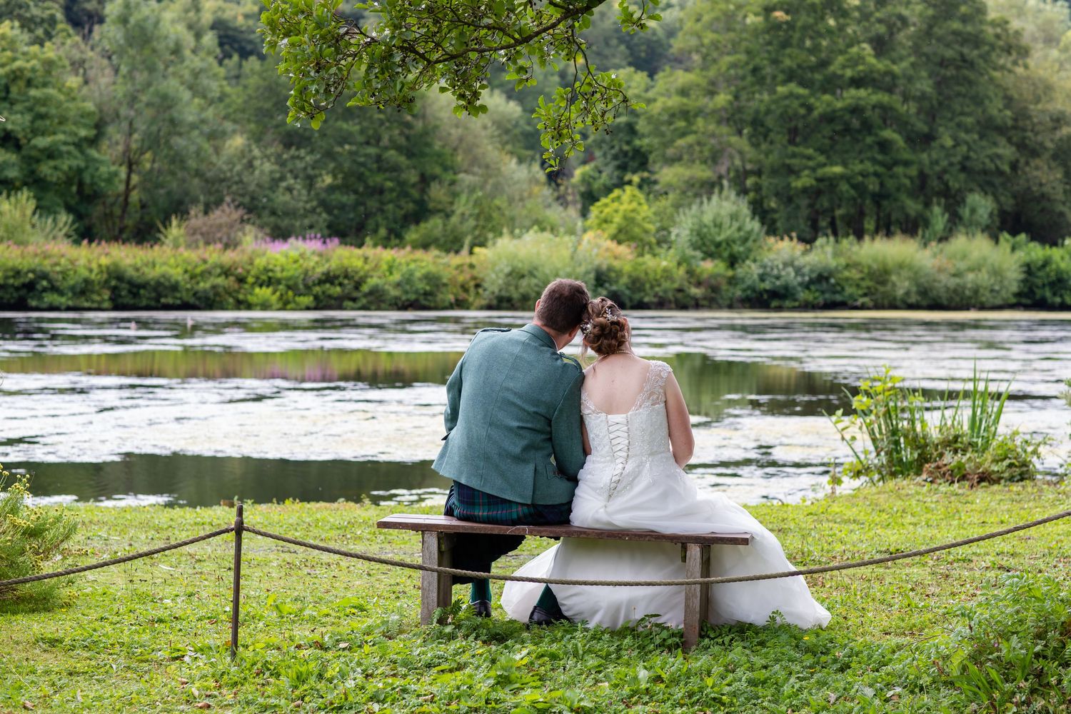 People sitting on benches near a serene lakeside setting surrounded by green trees.