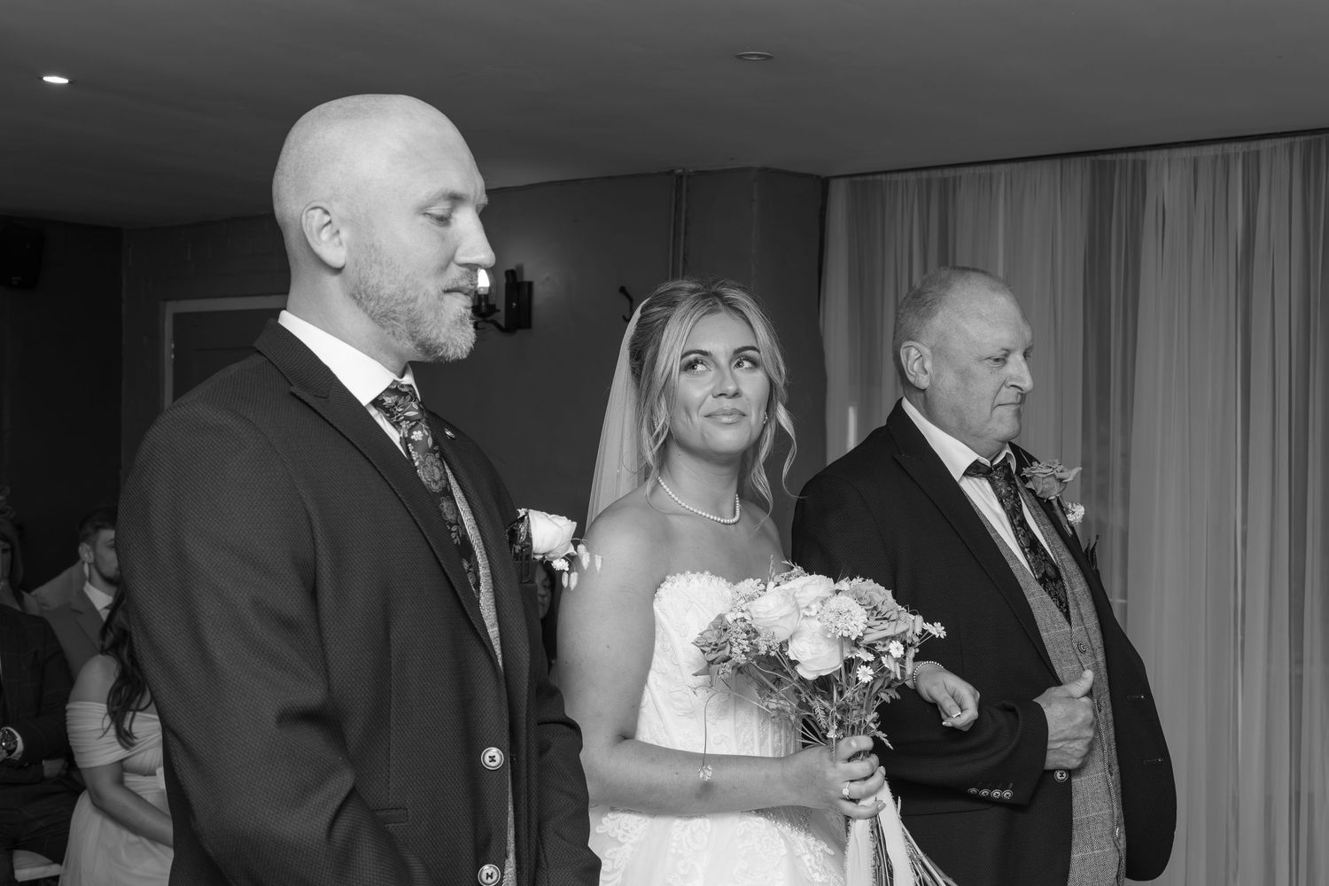 Black and white wedding ceremony photo of bride in veil holding bouquet standing between two men in suits.