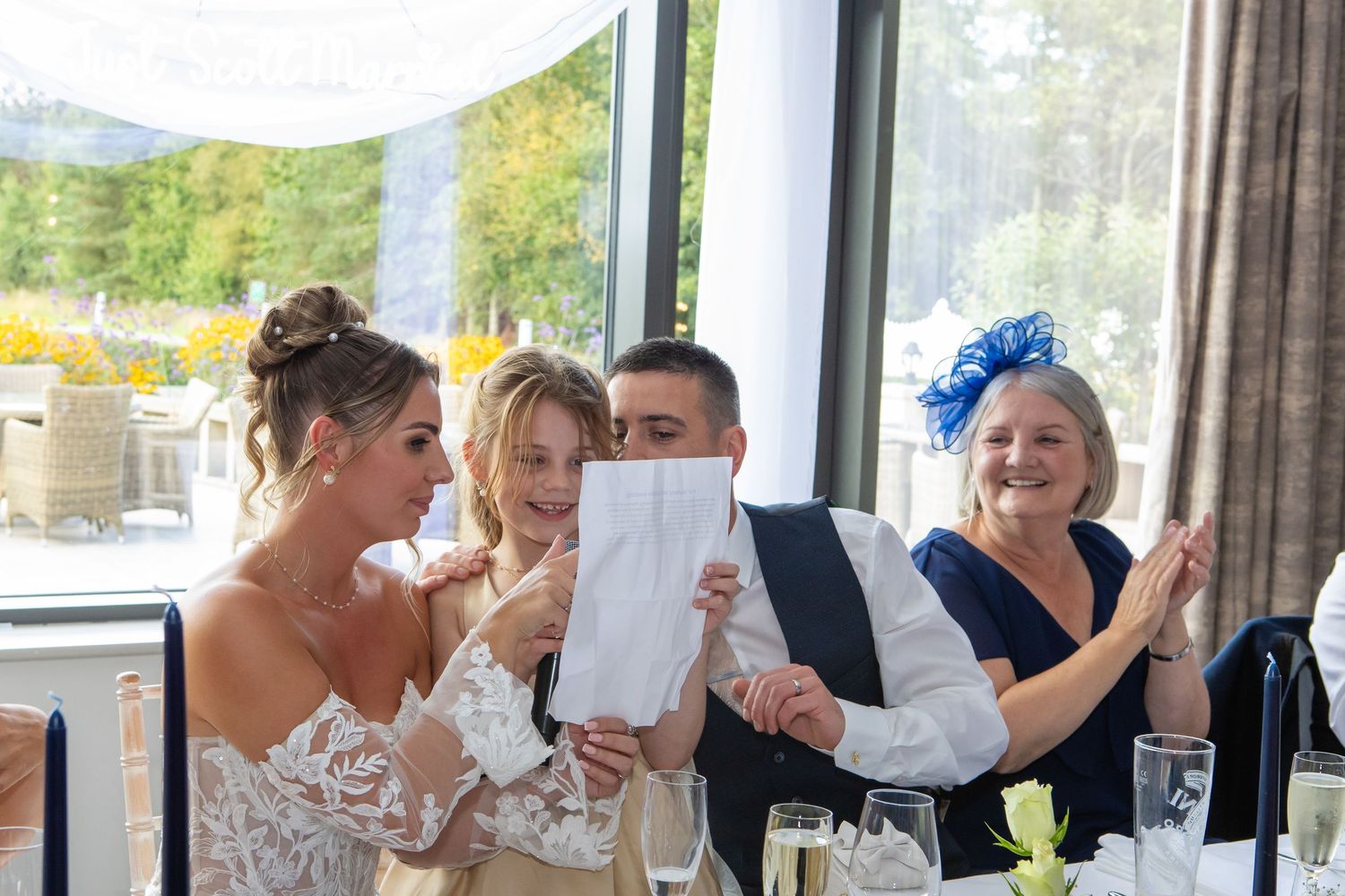 Wedding reception guests raise glasses in a toast while seated at long tables in an elegant venue.