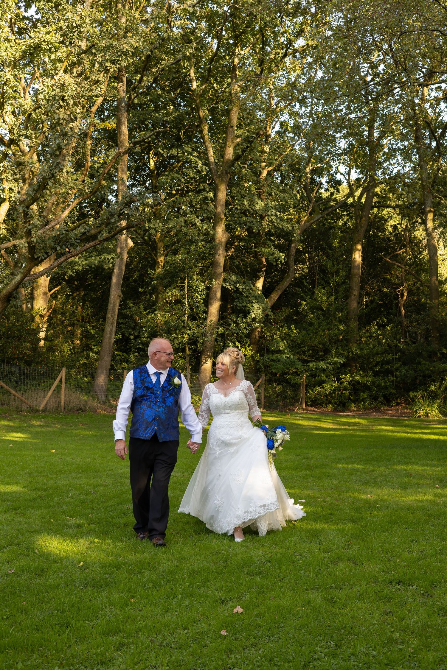 Bride and groom walking hand in hand across green lawn with trees in background on their wedding day.
