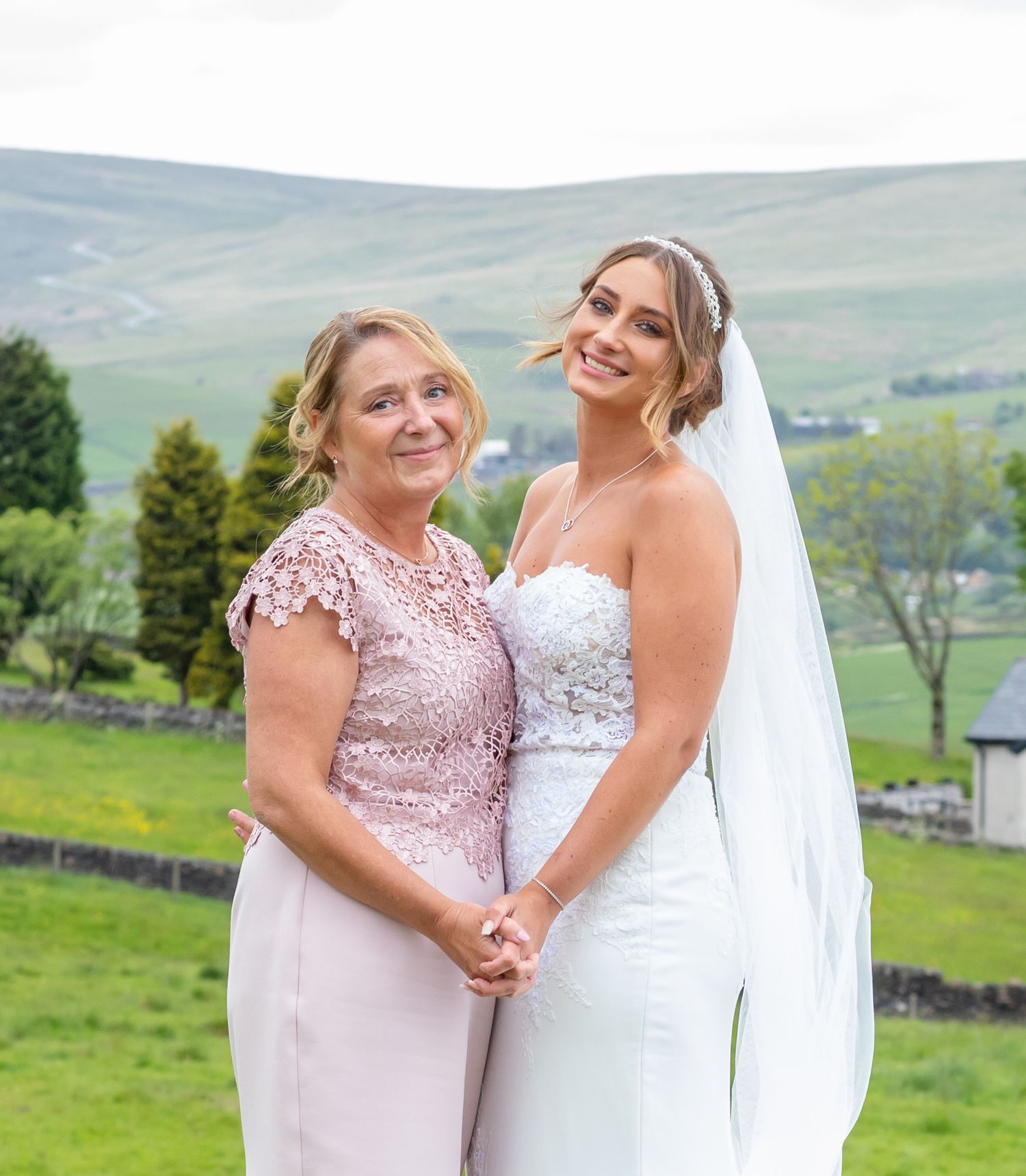Two people share an emotional embrace outdoors with rolling green hills and countryside in the background.