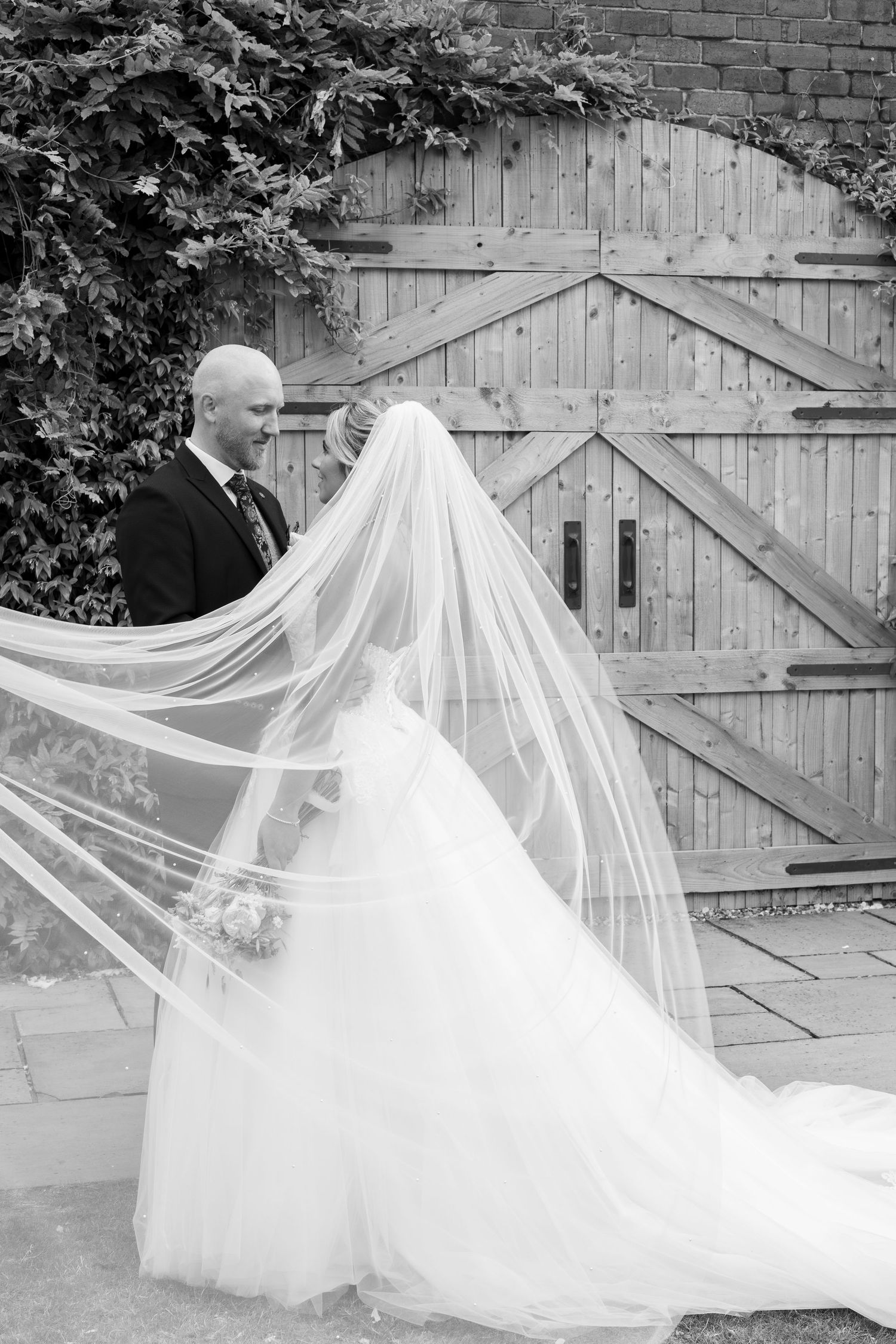Black and white wedding photo of bride in flowing veil and groom in suit standing before rustic wooden barn doors.