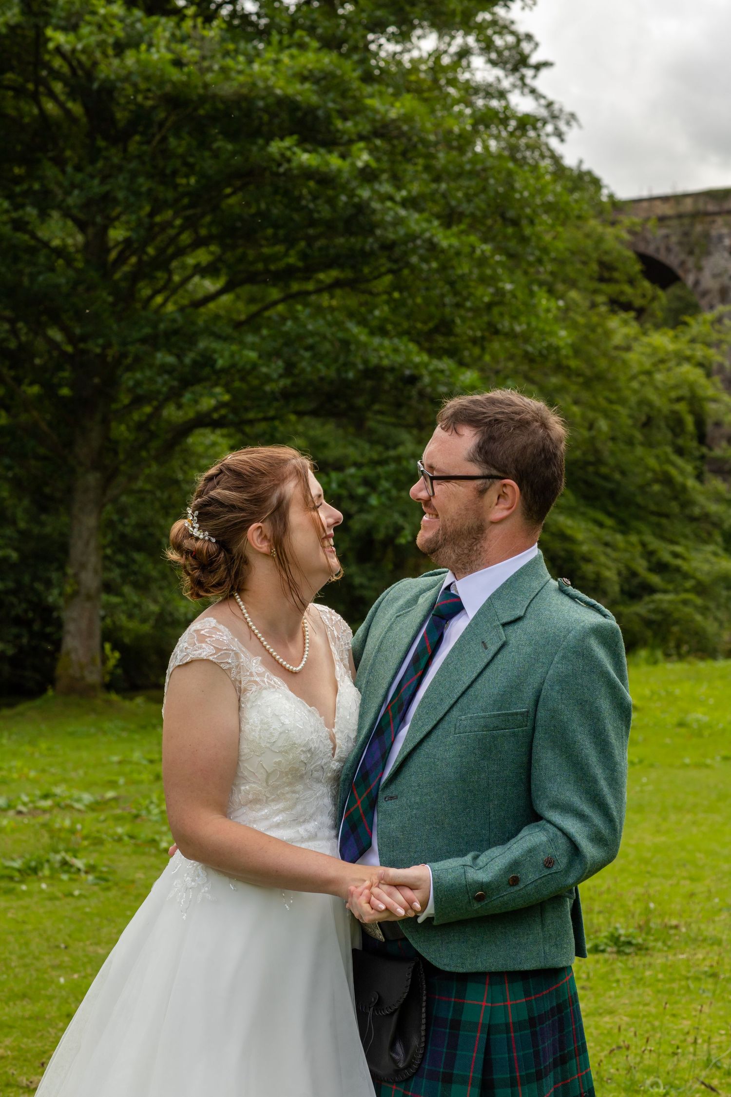A couple shares a romantic moment on their wedding day in a lush green garden, with the groom wearing a traditional kilt.