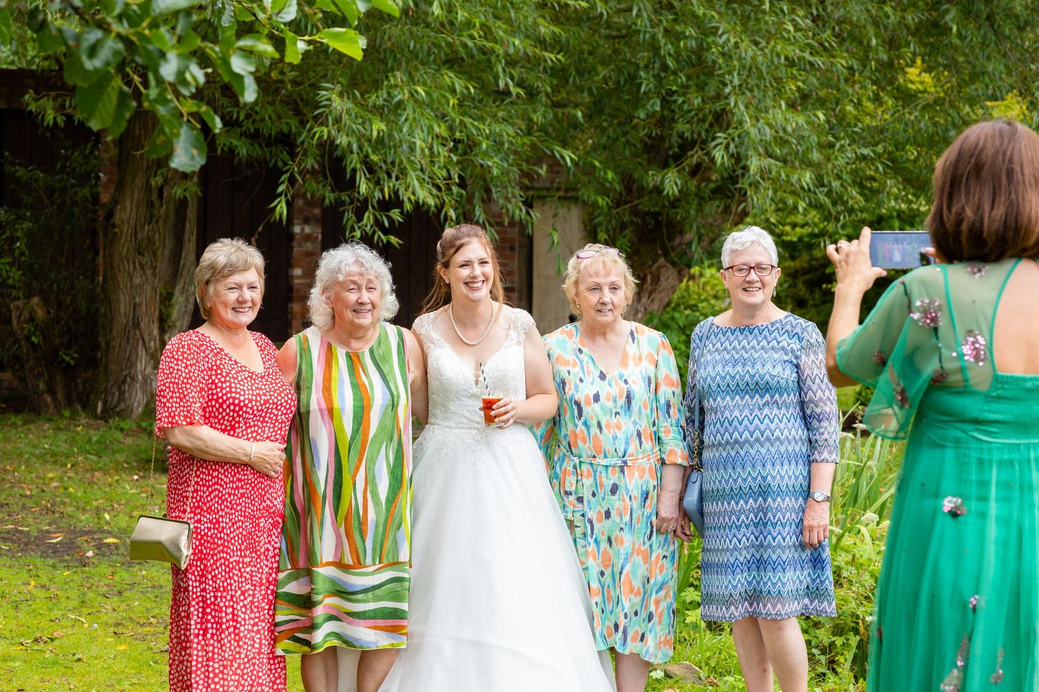 A group of women in colorful dresses and a bride in white gather outdoors on a green lawn for photos.