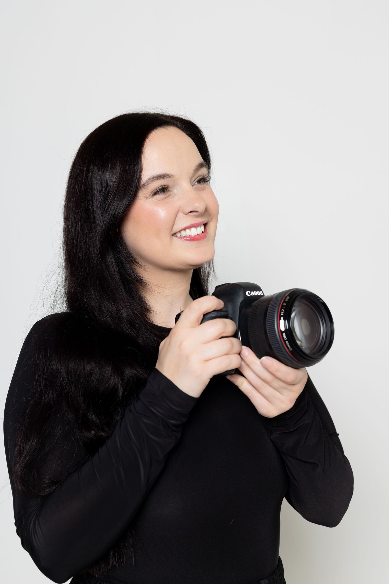 Photographer in black sweater poses with camera against white background.