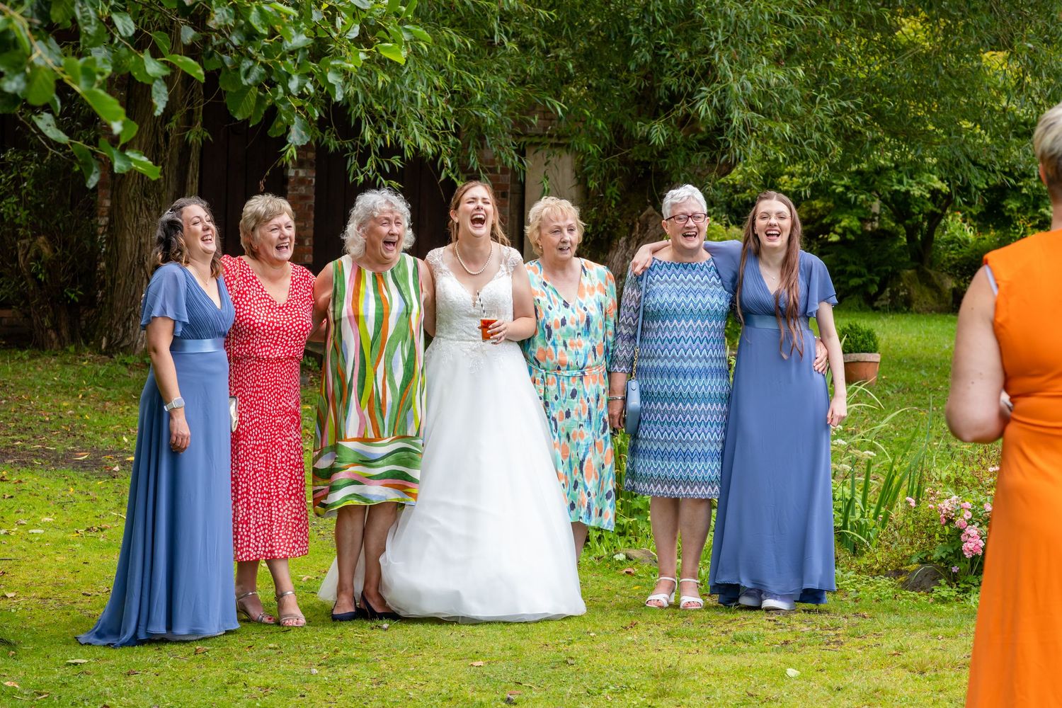 Wedding party members gather outdoors in colorful blue, orange, and pink dresses for group photos in a garden setting.