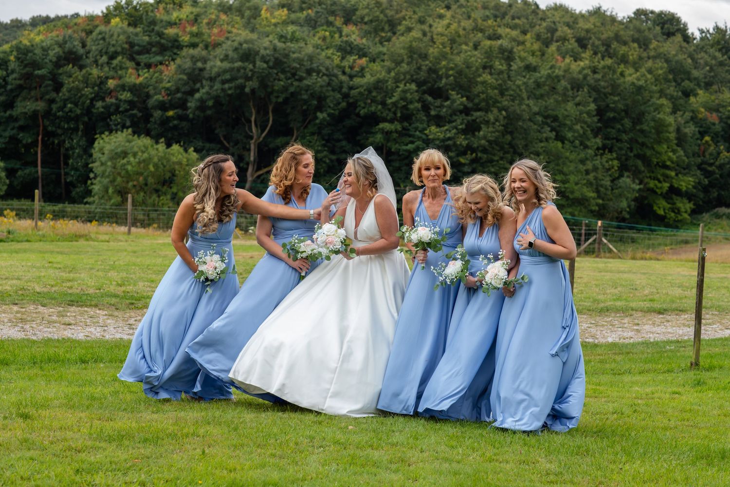 Wedding party members in light blue dresses pose together on a grassy hillside.