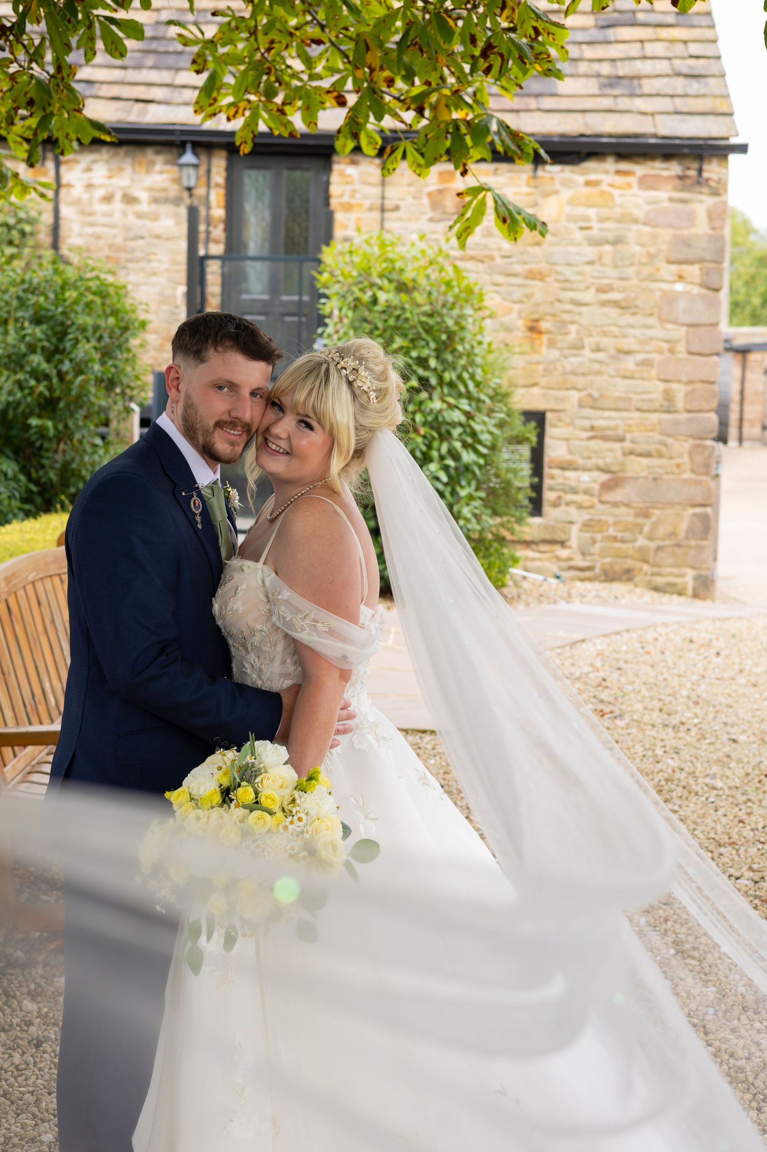 A bride and groom embrace outdoors by a stone building, her veil flowing as she holds a yellow and white bouquet.