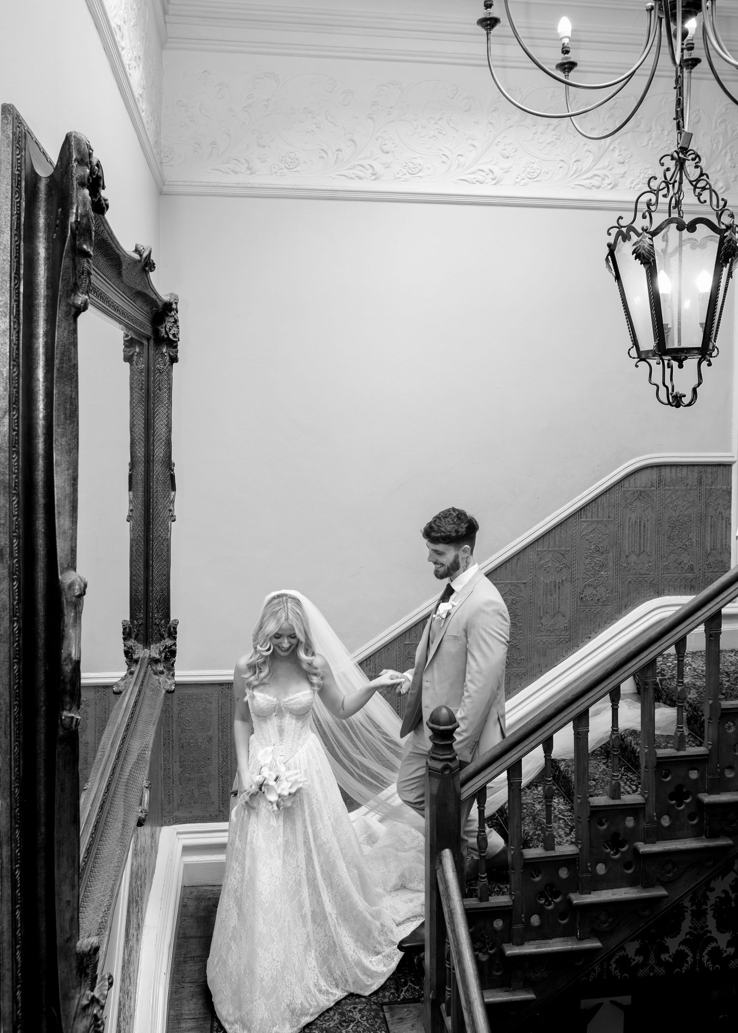 Groom lifting bride's veil on elegant staircase in black and white wedding portrait with chandelier.