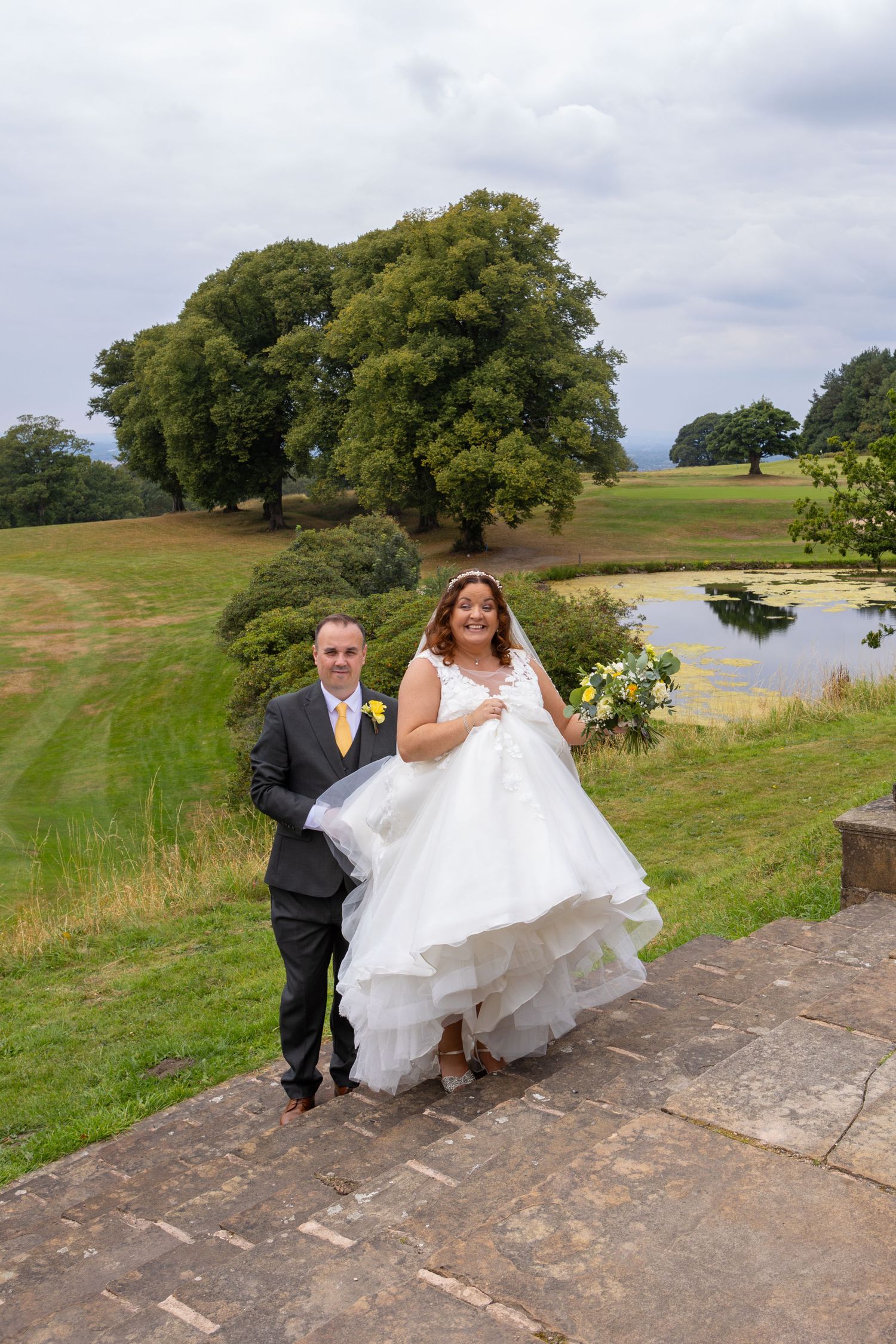 Wedding couple stands on rustic stone path overlooking scenic countryside with pond.