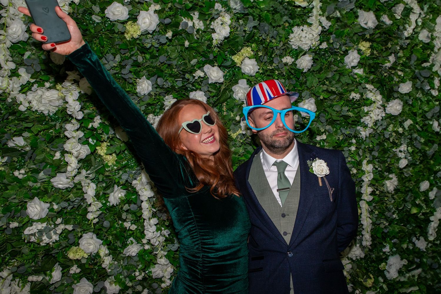Two people pose playfully against a white floral wall backdrop wearing sunglasses and formal attire.
