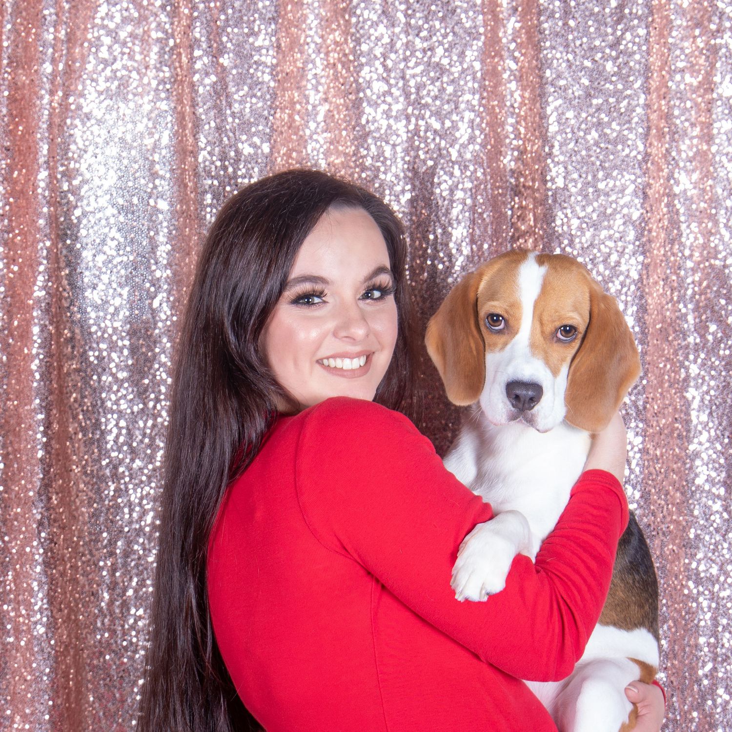 Portrait of a photographer in red sweater holding a cute beagle dog against a sparkly pink backdrop.