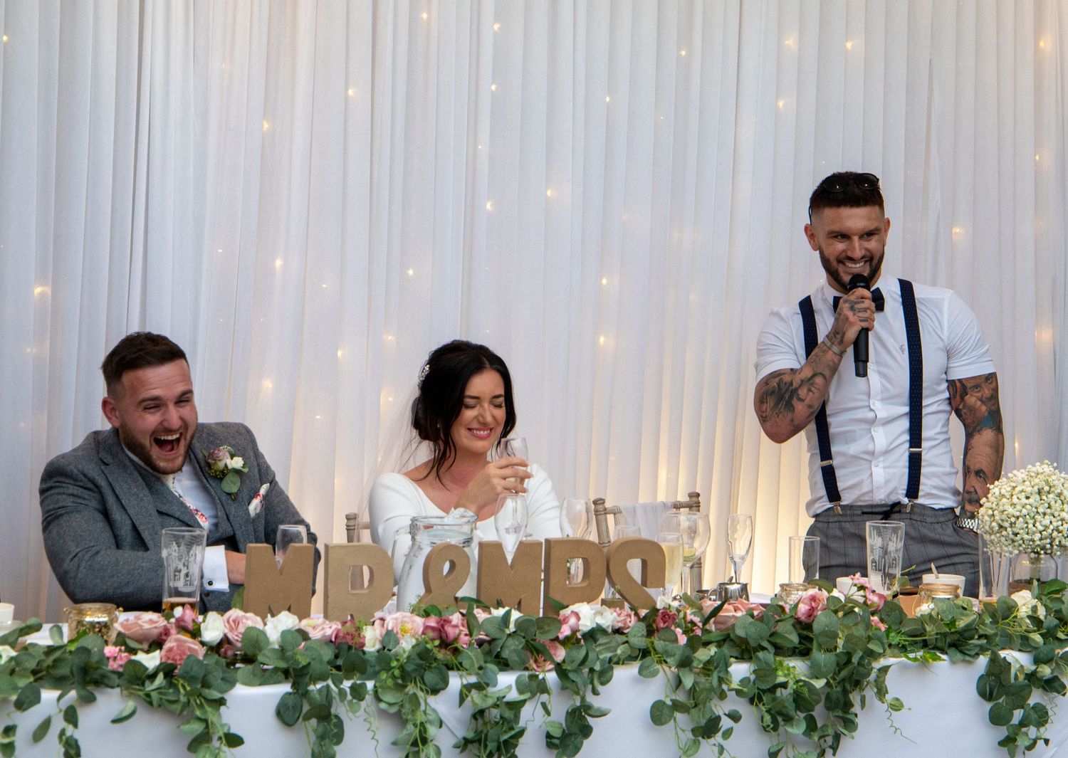 Wedding reception head table decorated with fairy lights and greenery garland against white backdrop.