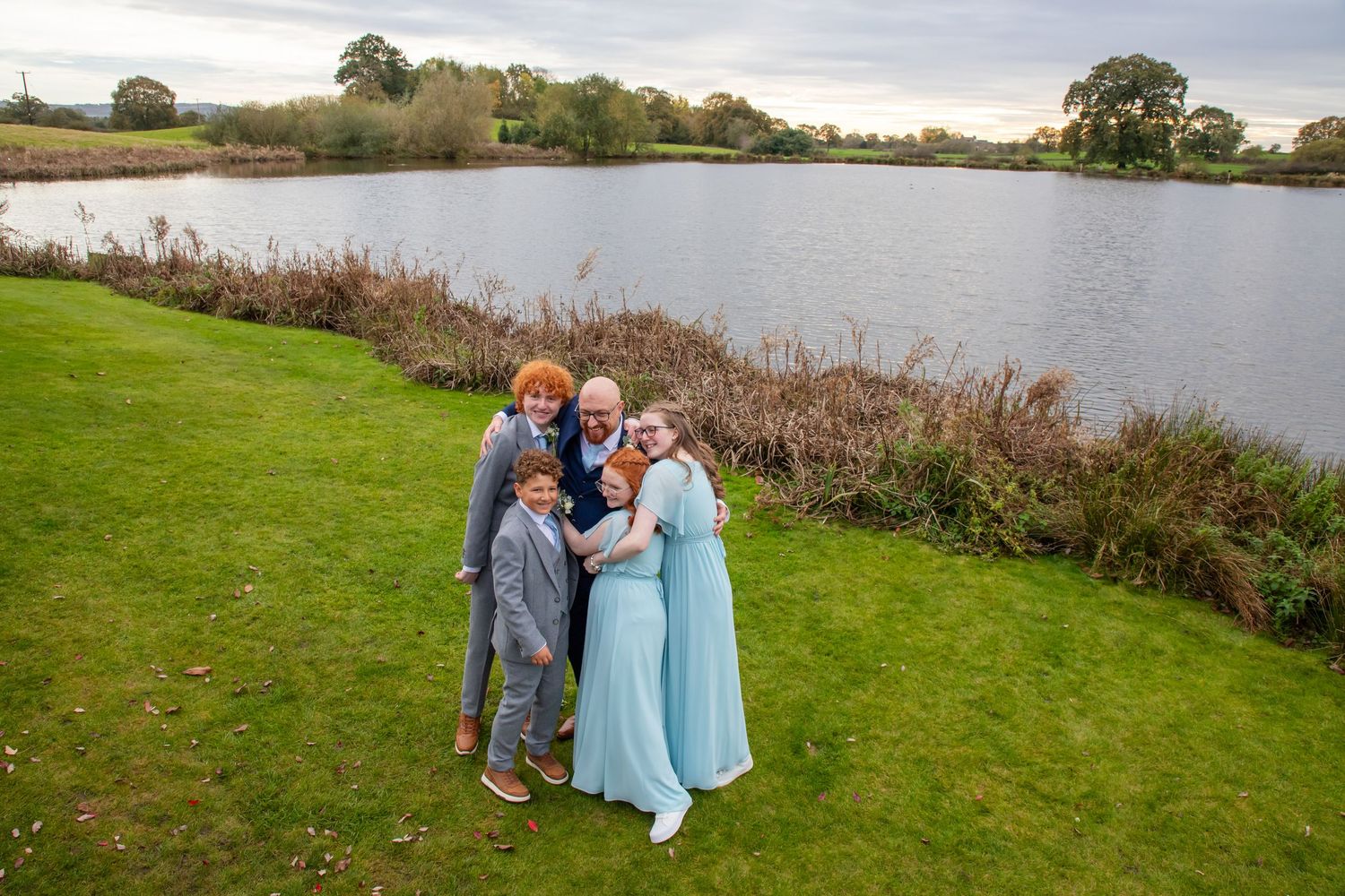 A scenic lakeside photo session on green grass with water and trees in the background during golden hour.