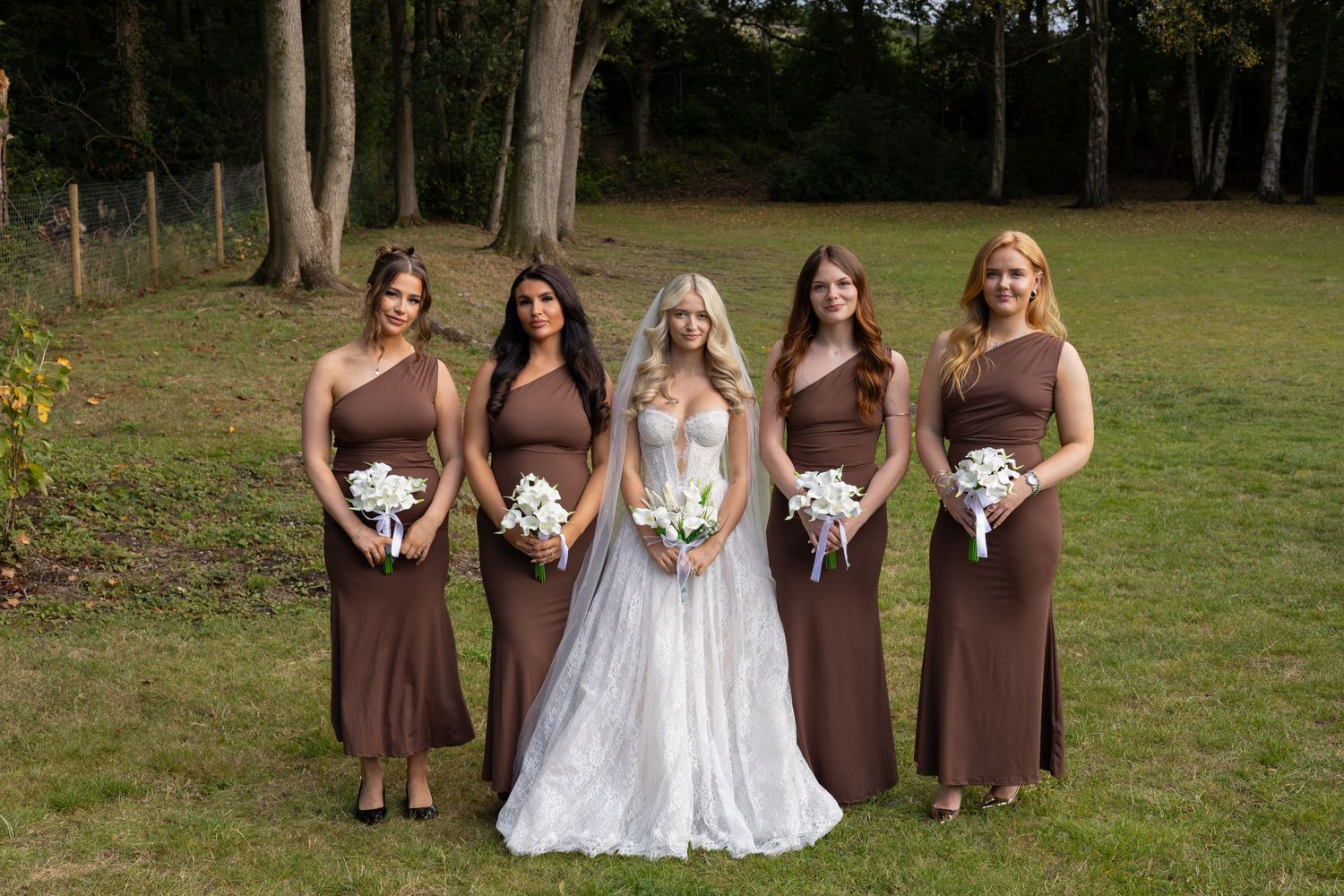 Bride in white gown with three bridesmaids in brown dresses holding bouquets outdoors on green lawn with trees.