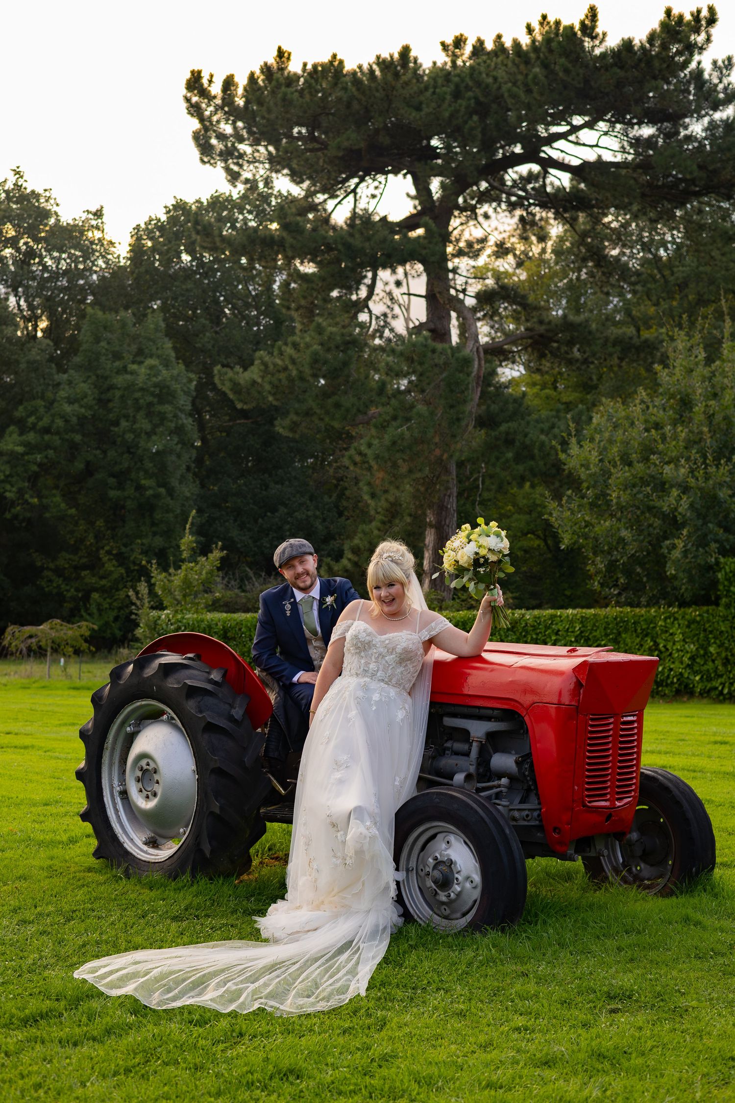 Bride in white gown and groom in suit pose with a vintage red tractor on green lawn surrounded by tall trees.