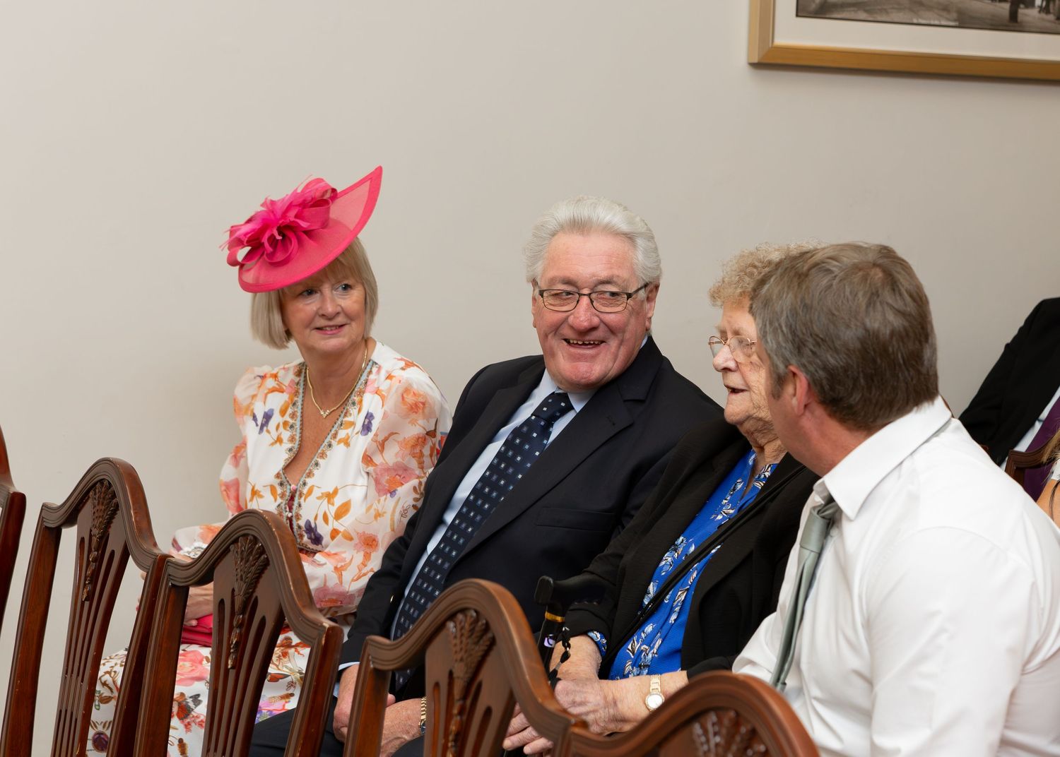 Elegantly dressed group conversing at formal event, woman in pink floral hat and dress seated with suited man.
