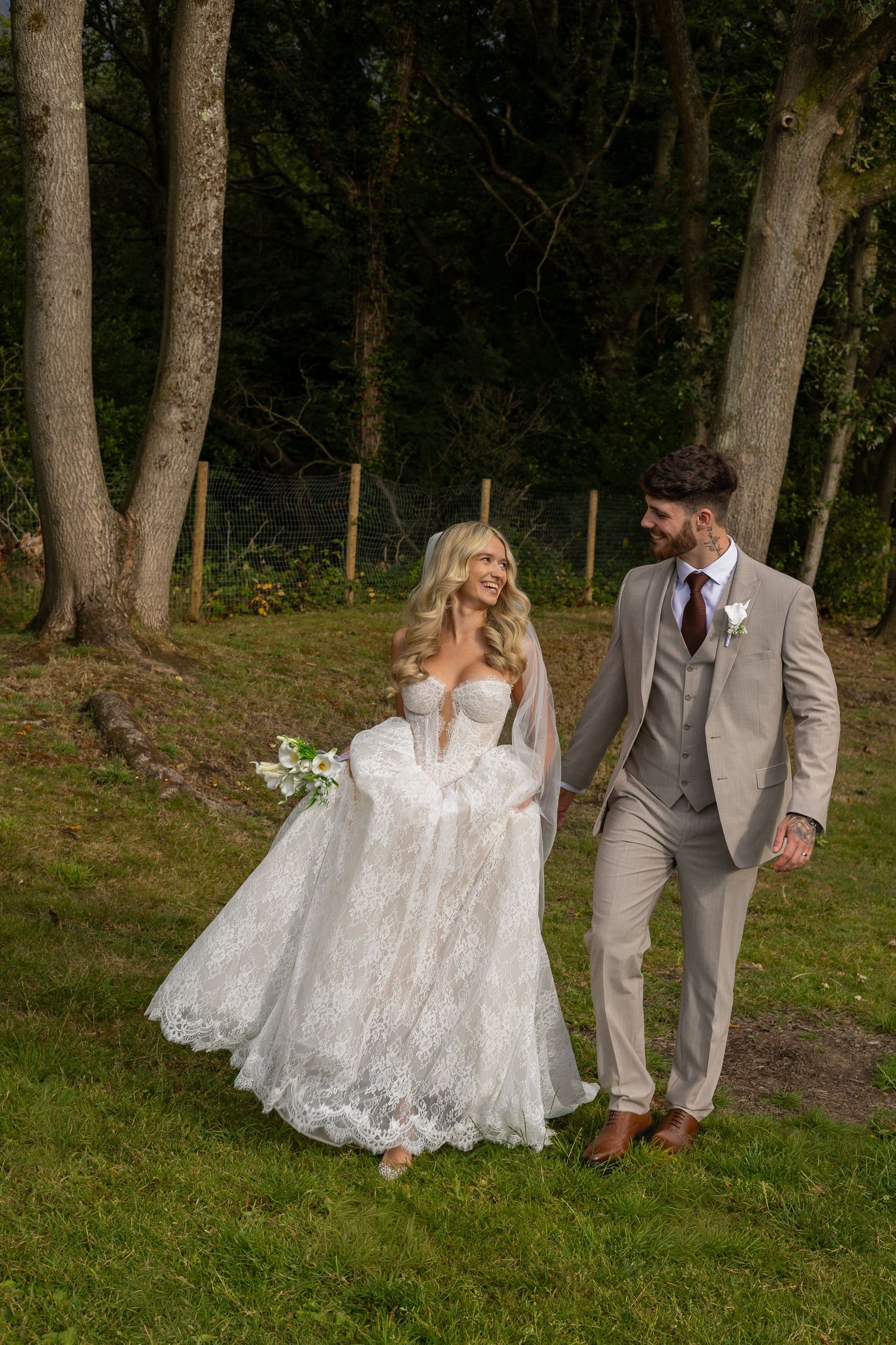 Bride and groom walk hand in hand through lush green woodland, she in lace gown, he in tan suit.