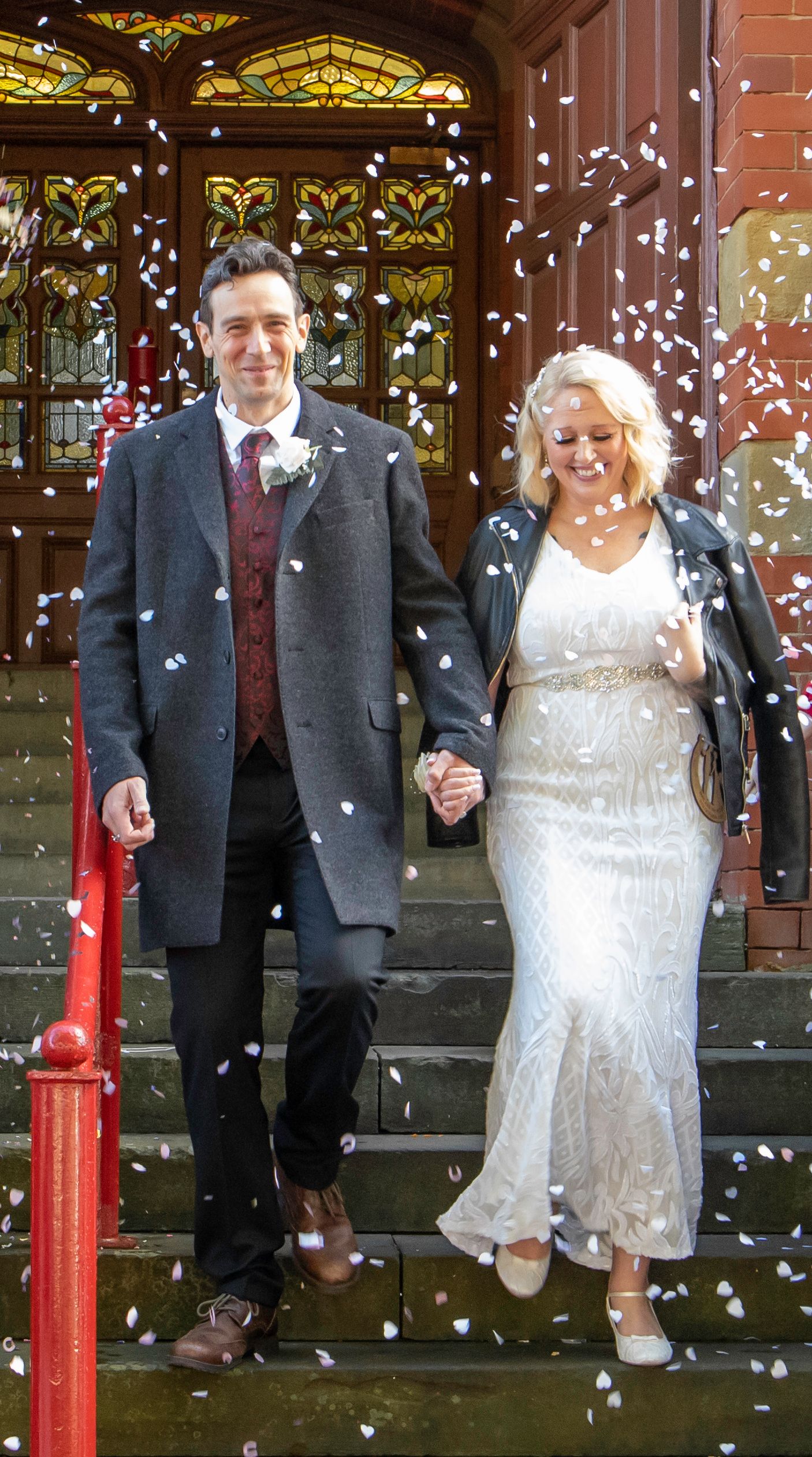 A joyful wedding couple being showered with confetti on brick steps with stained glass windows visible.