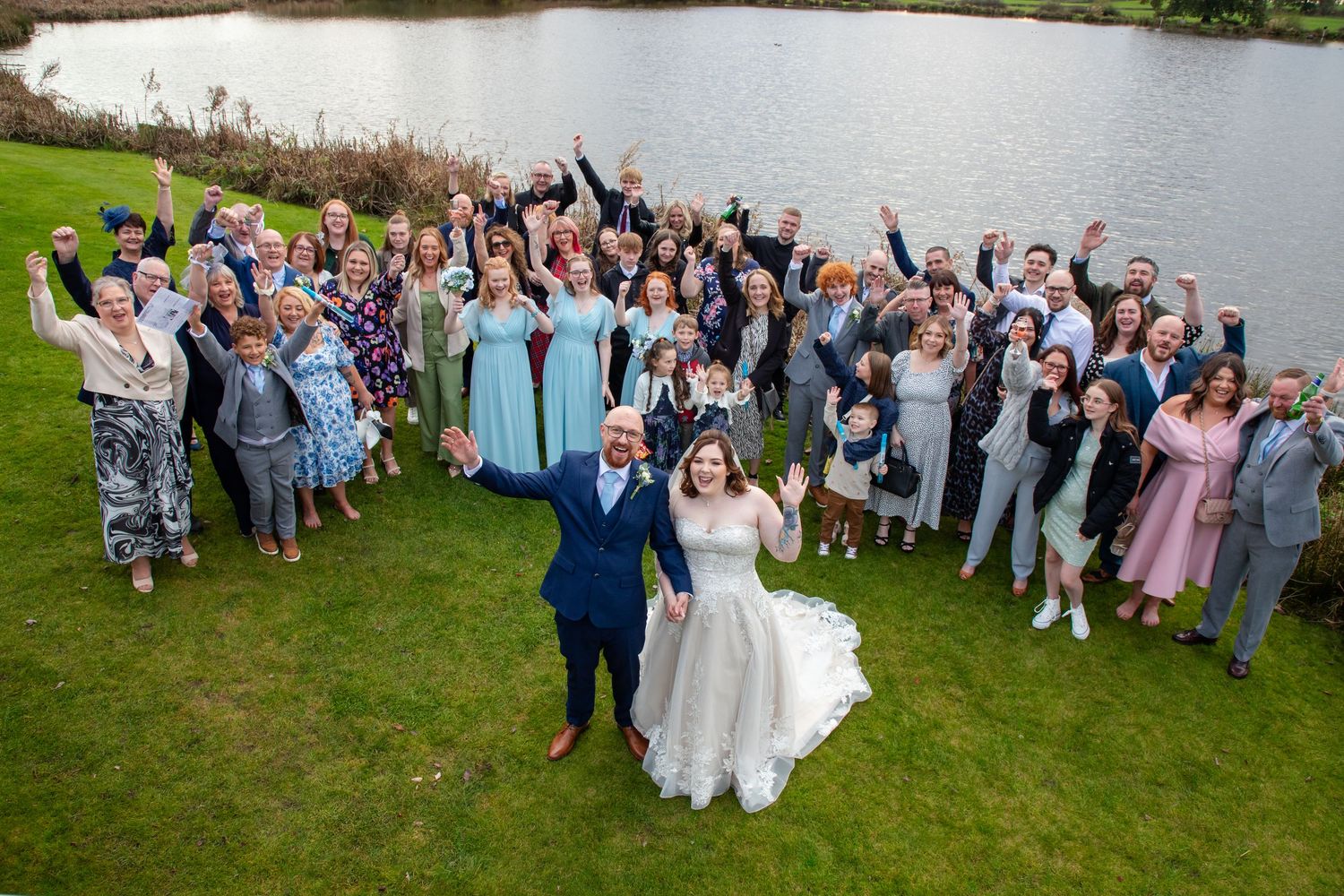 Large wedding party poses for group photos on a grassy lawn beside a lake during an outdoor celebration.